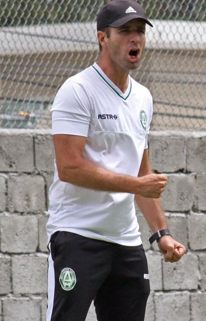 A man in sports attire passionately cheering, wearing a cap and clenching his fist in front of a chain-link fence.