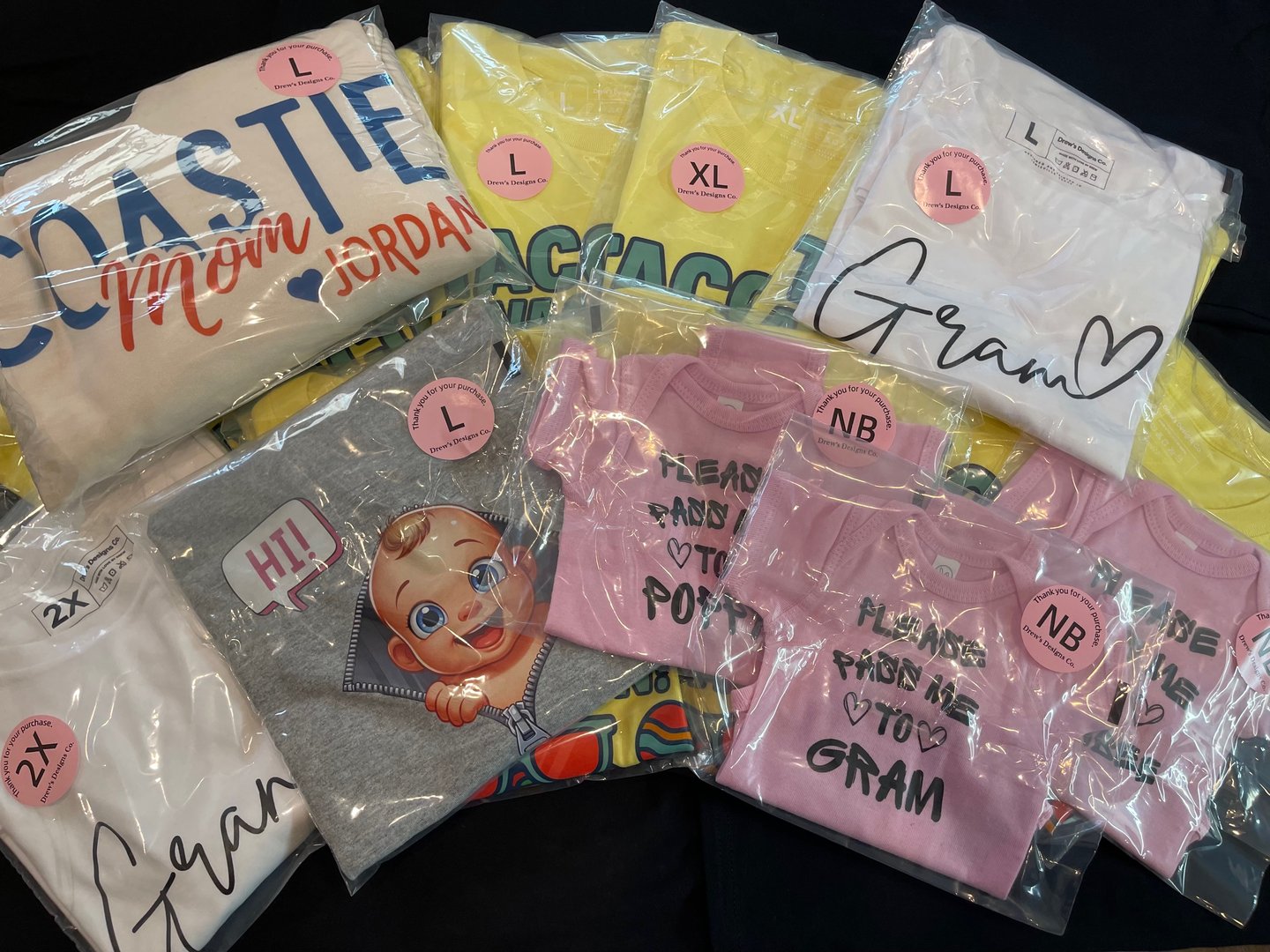 A variety of folded t-shirts in plastic bags, labeled with sizes and featuring graphics and text like Gram and Coastie Mom.