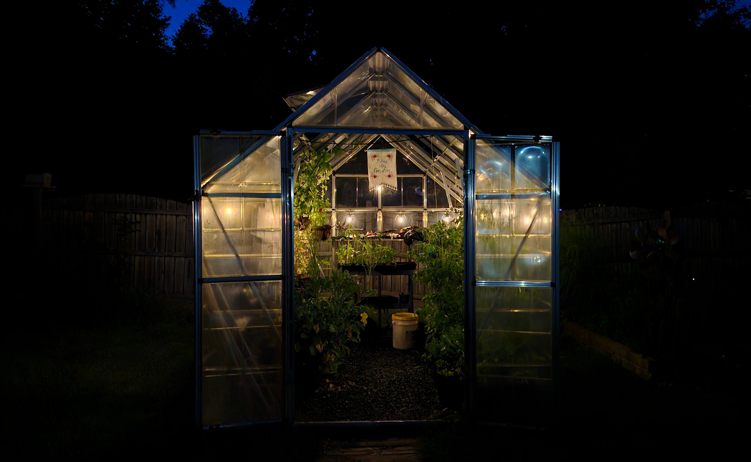A serene nighttime view of a backyard greenhouse lit from within, showcasing lush greenery and a warm, inviting atmosphere perfect for homesteading or gardening themes.