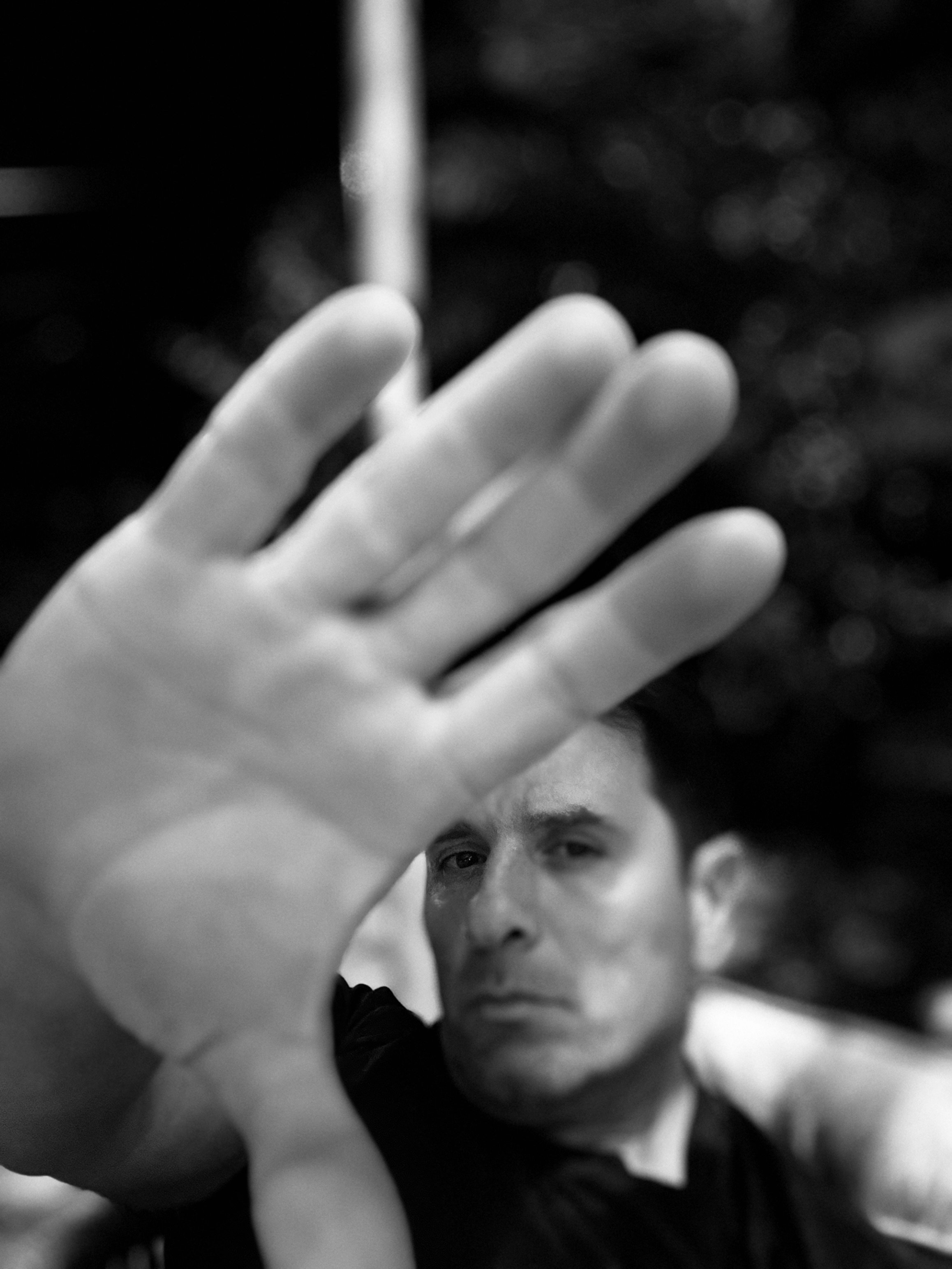 Black and white photo of a man raising his hand towards the camera, partially obscuring his face with a defocused background.