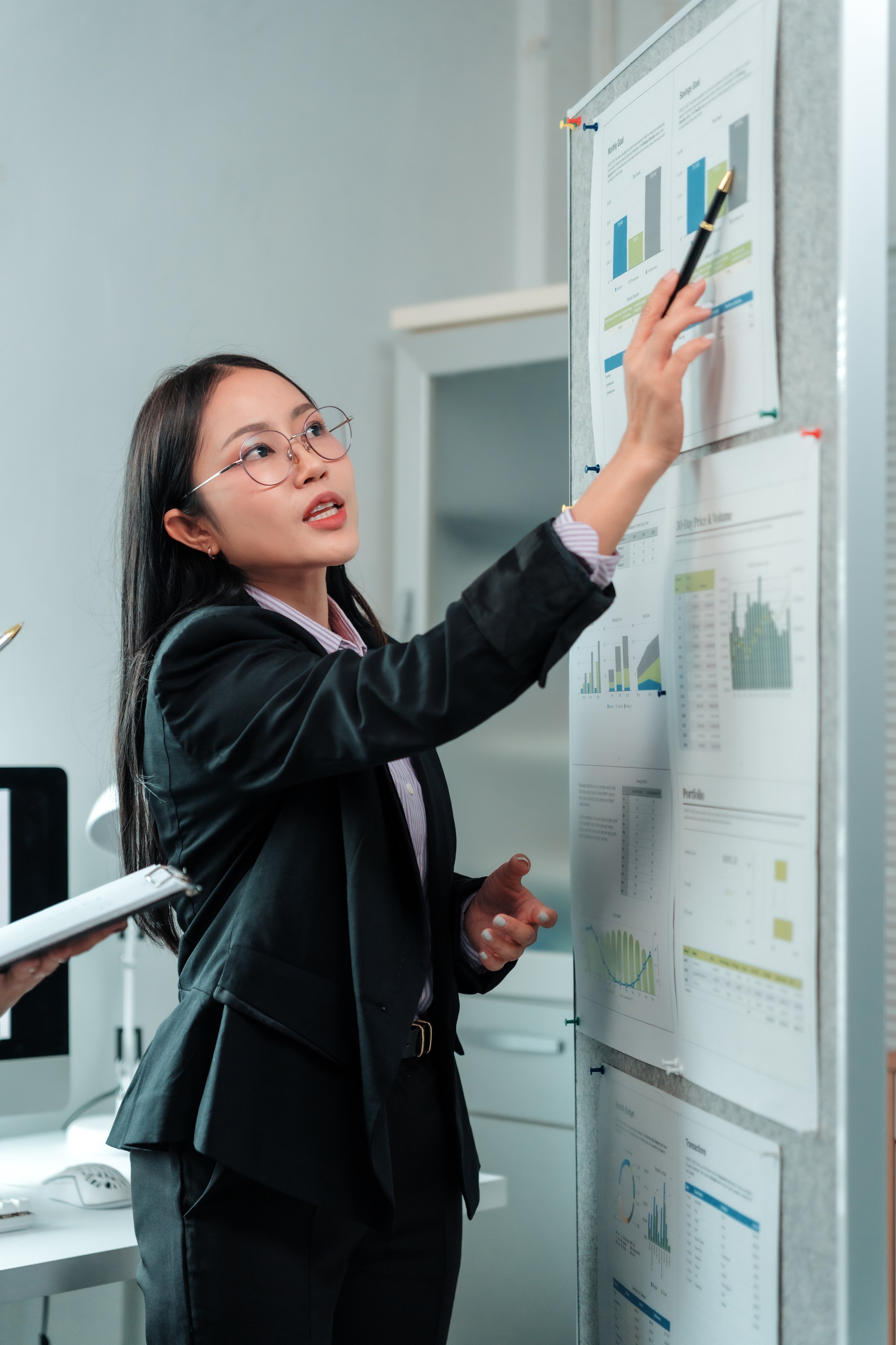 Young Asian businesswoman wearing glasses and a suit pointing at charts on a whiteboard during a presentation in a modern office, explaining data analysis and business strategies