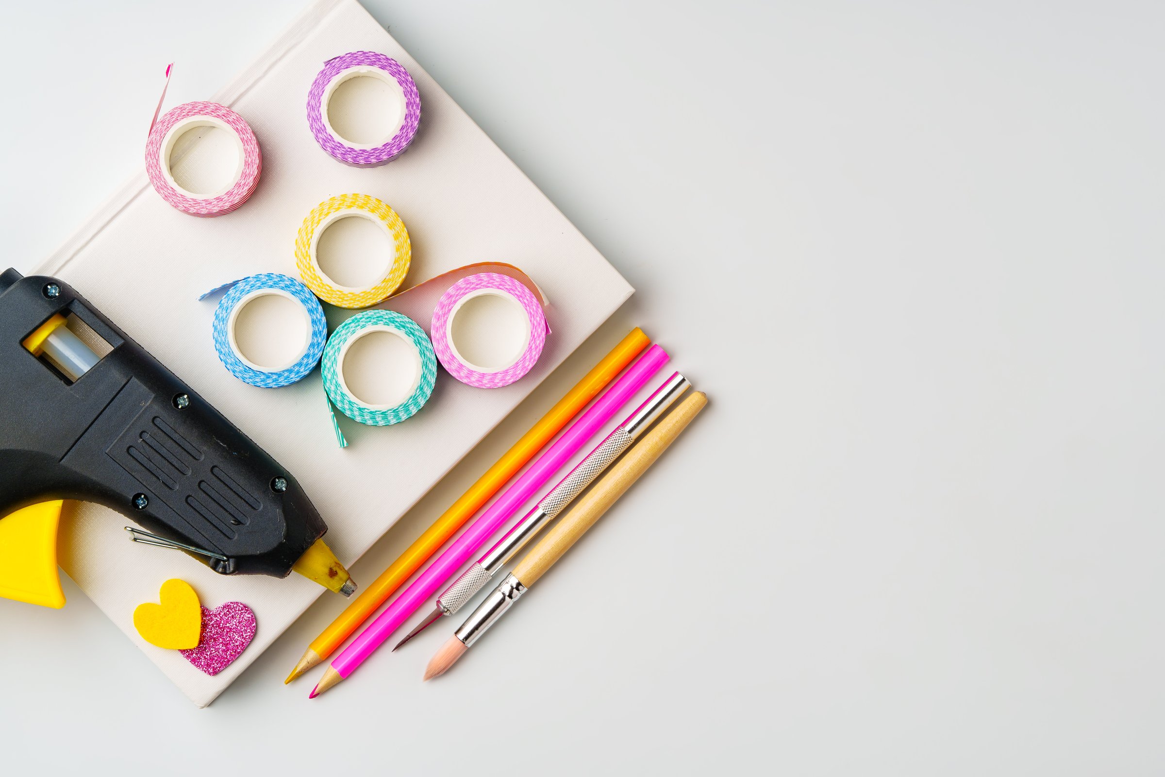 Various crafting tools are neatly arranged on a white surface, featuring a glue gun, rolls of decorative tape in vibrant colors, and colored pencils ready for creative use.
