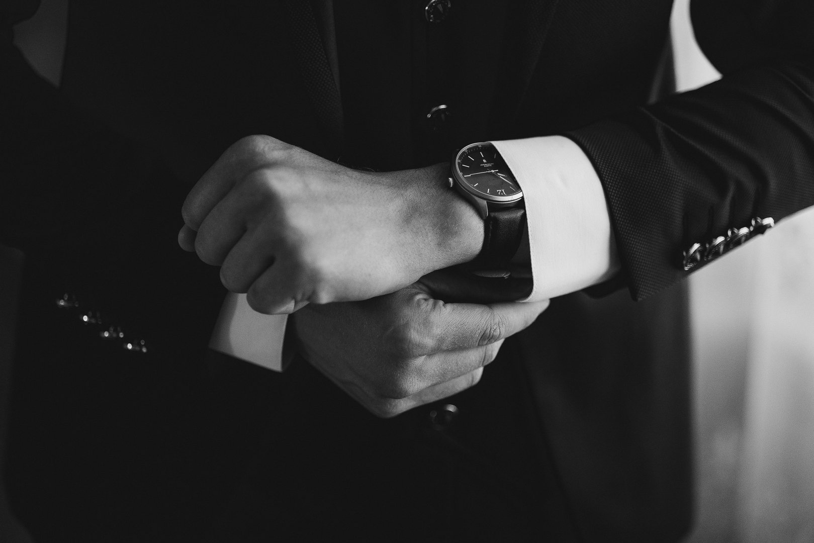 A man is wearing a black suit and is looking at his watch. The watch is black and has a silver band. The man is in a hurry or is checking the time