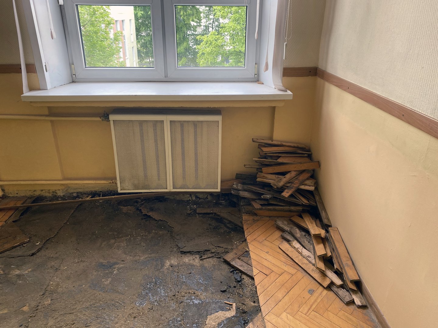 Stack of removed wooden parquet planks piled in corner of empty room with damaged floor during renovation near window.