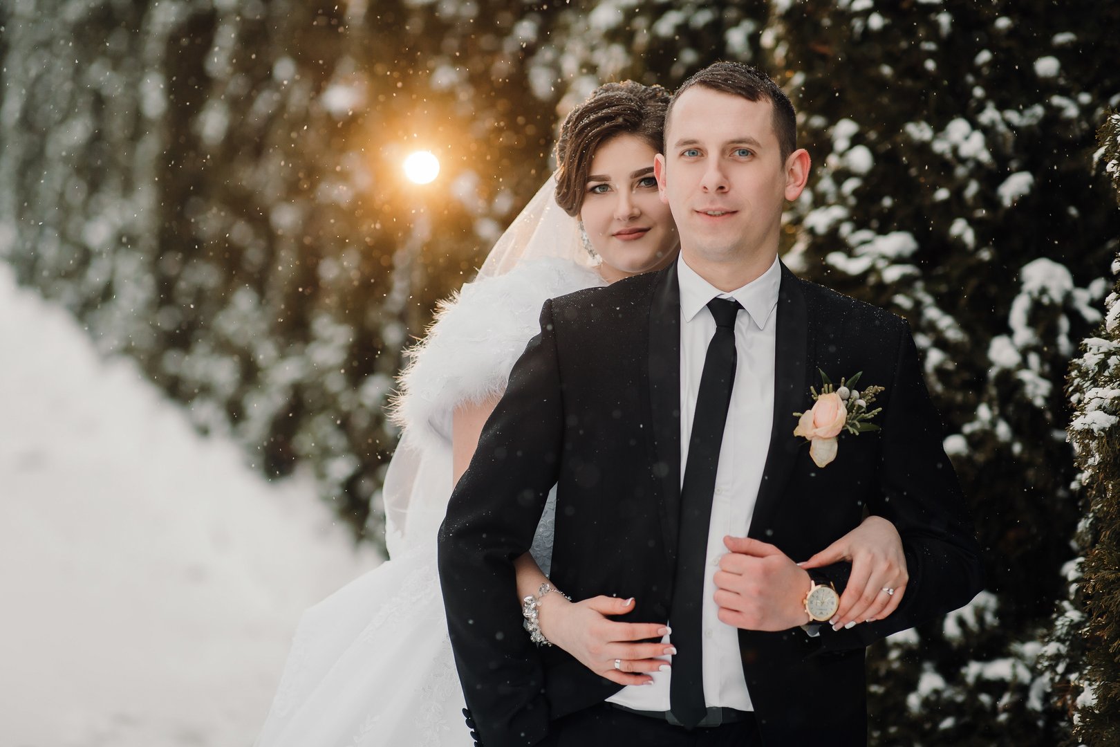 A bride and groom are posing for a picture in the snow. The bride is wearing a white dress and a fur stole, while the groom is wearing a black suit and a tie. The couple is standing in front of a tree