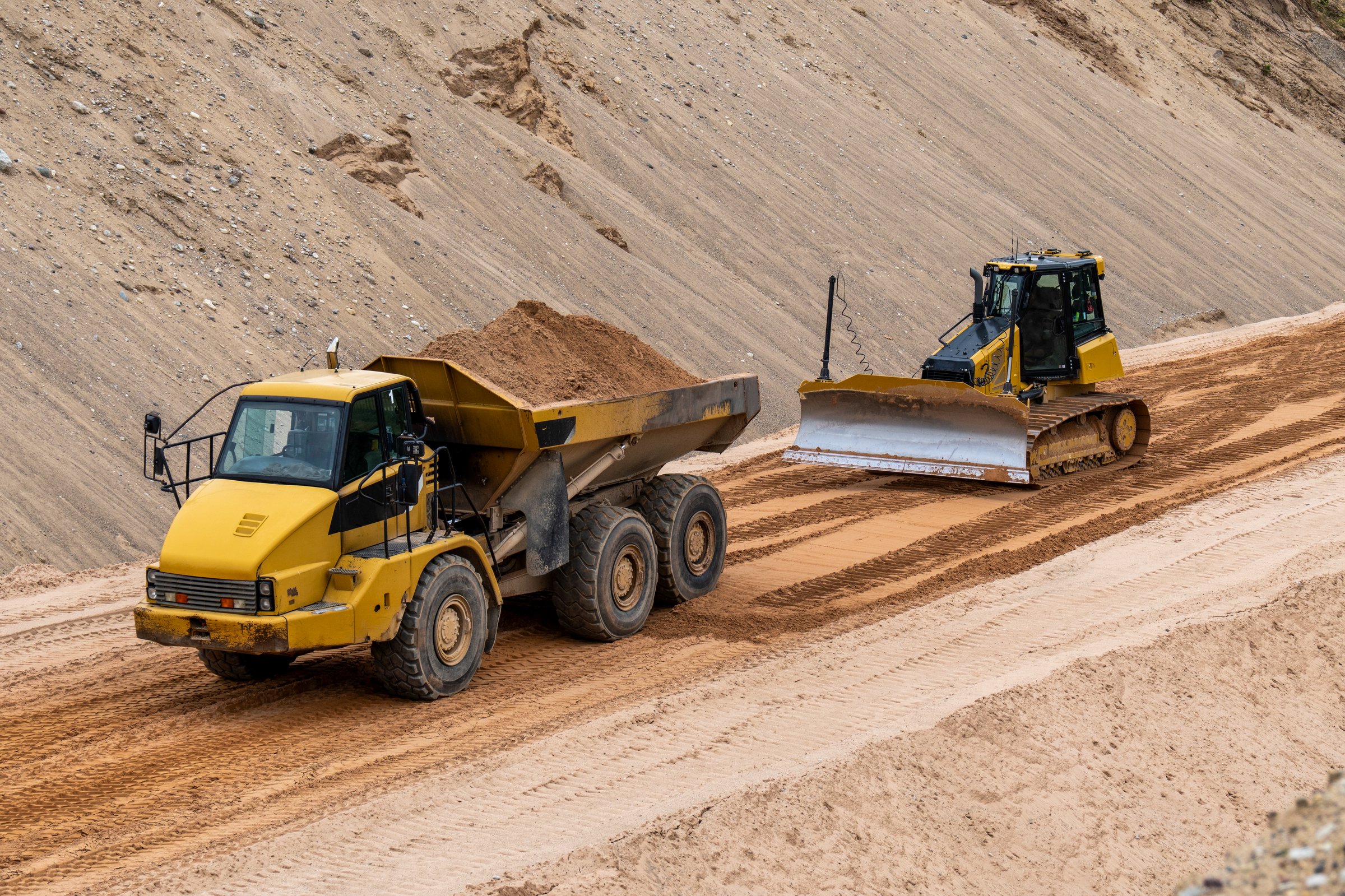 Heavy construction vehicles working on sandy site, dump truck and bulldozer leveling ground at quarry or infrastructure construction zone