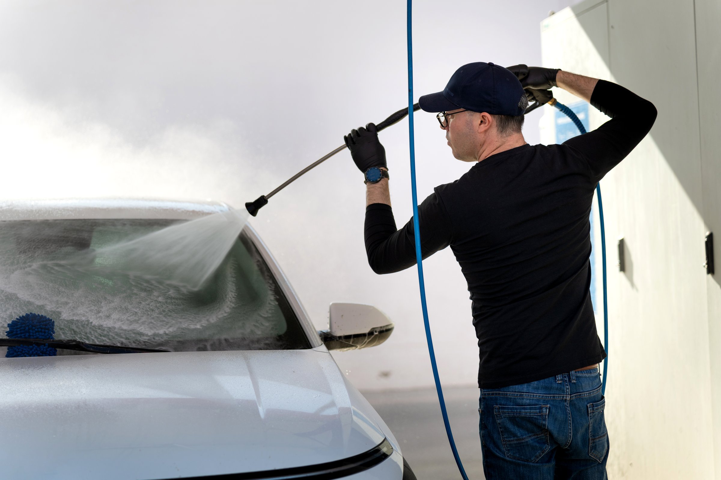 Man in workwear cleaning a white car with a high-pressure water sprayer, offering professional detailing service