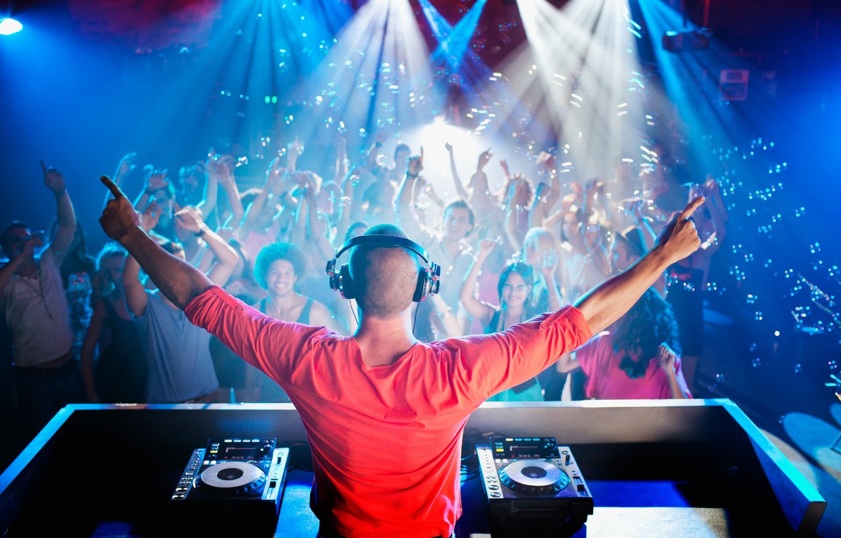 DJ in red shirt performing on stage with a lively crowd dancing under blue and white lights in a club setting.