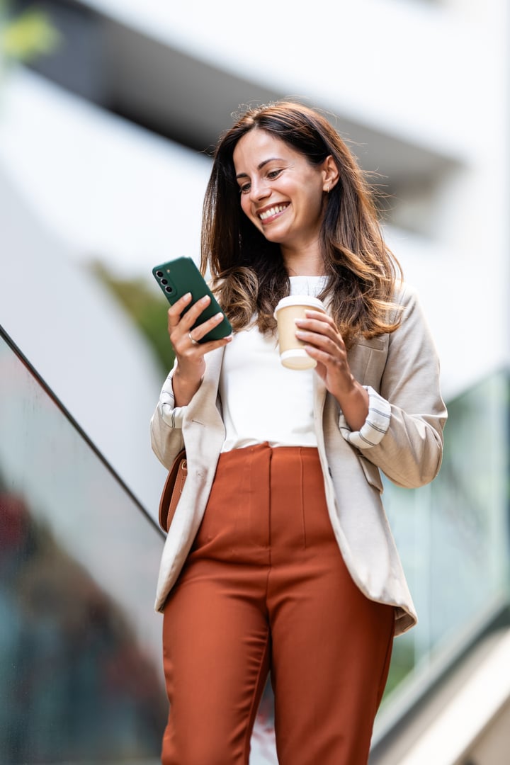 Happy businesswoman walking outdoors, sipping takeaway coffee while engaging with her smartphone, enjoying a moment of urban lifestyle