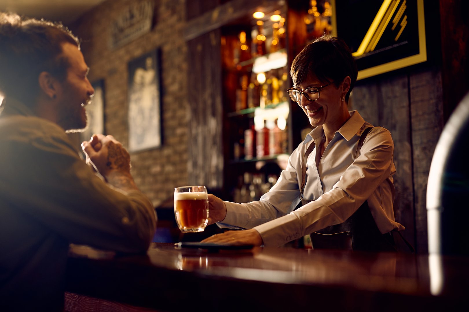 Happy waitress serving beer to a customer at bar counter.