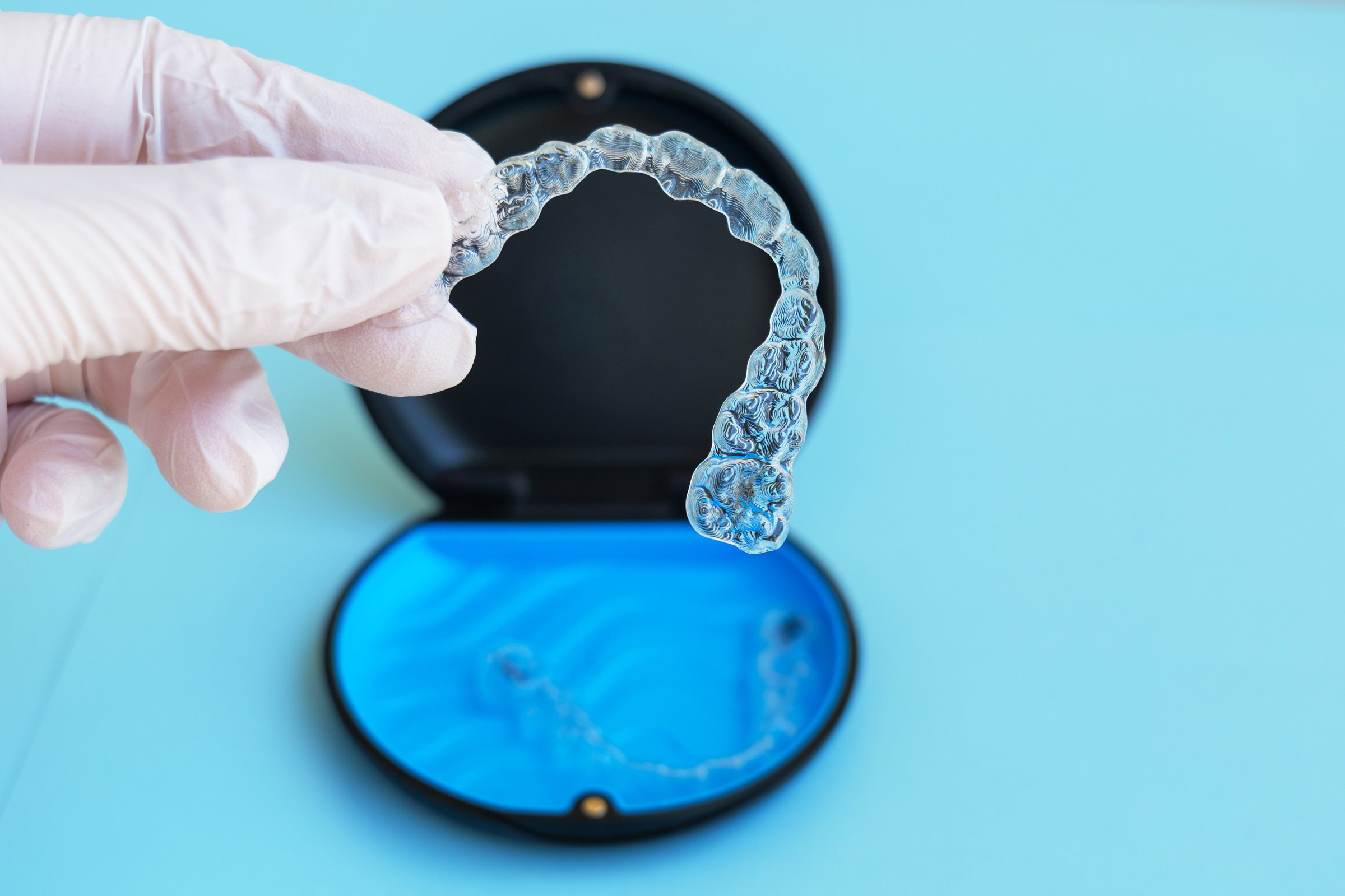 Gloved hand holding a clear orthodontic aligner with the storage case visible in the background, highlighting precision, cleanliness and modern dental care tools in a minimal clinical setting