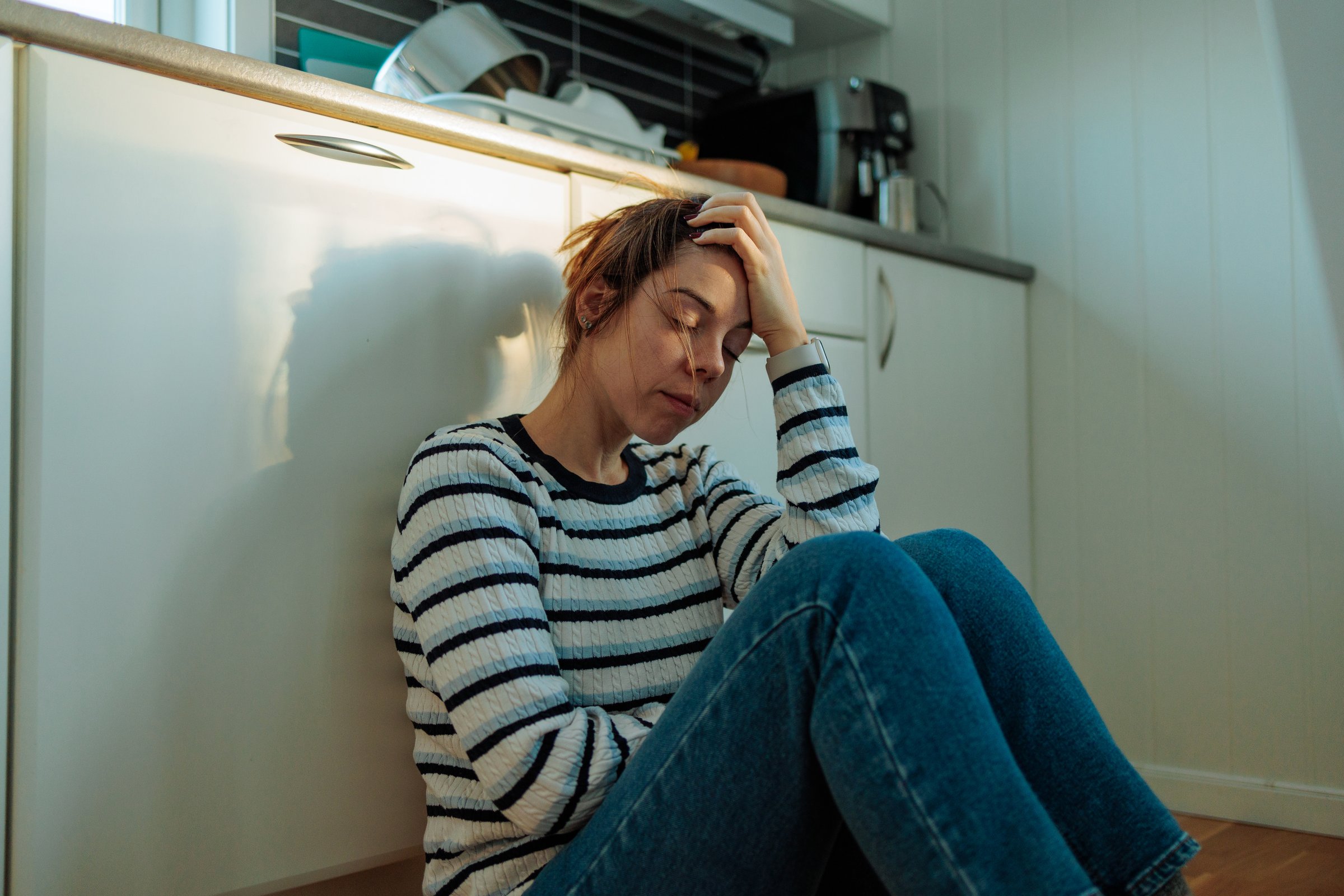 Young woman sitting on the kitchen floor with her head in her hand, suffering from depression, loneliness, and mental health issues