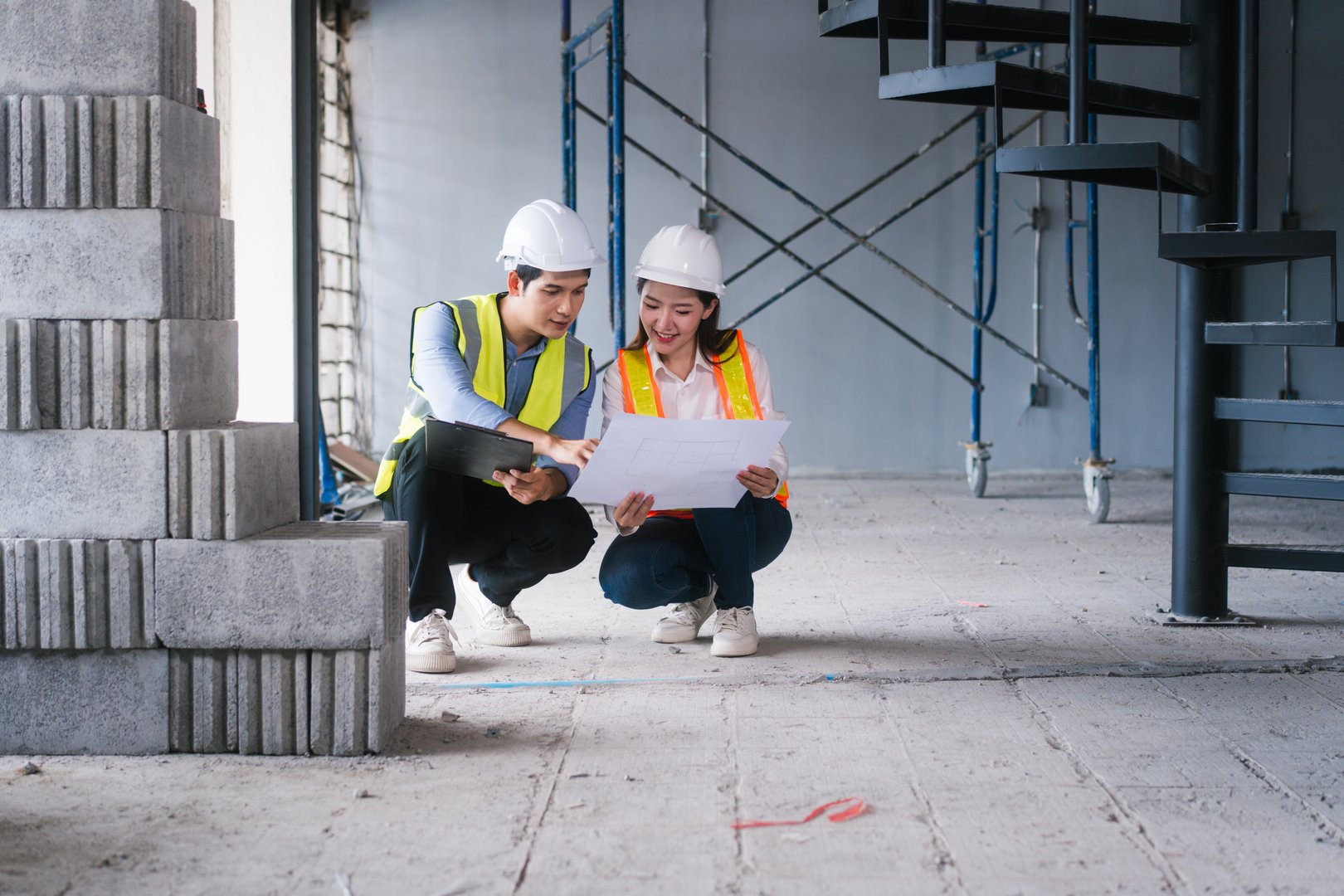 Asian male and female engineers wearing safety vests and helmets inspect structural elements, review blueprints, plan material choices bricks, steel, concrete during home renovation project on-site.