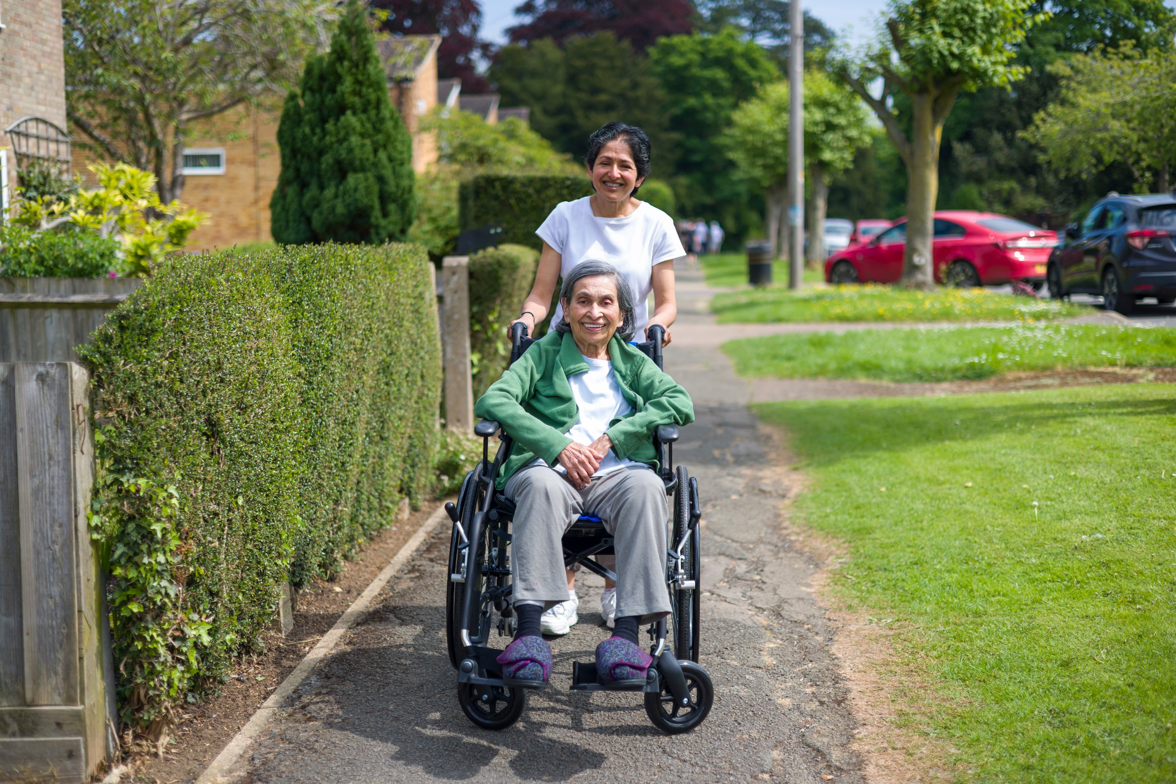 Old Indian woman in wheelchair outdoors on a pavement in a suburban street in summer, UK. Mother and daughter: may also depict a carer, care in the community