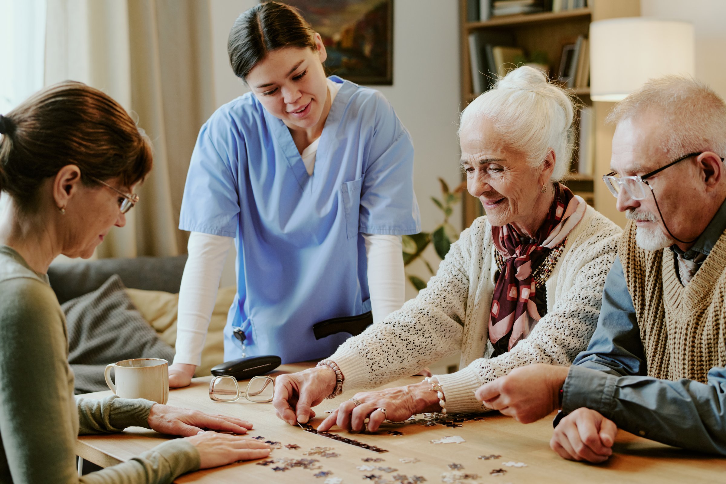 Senior Caucasian woman and senior Caucasian man assembling puzzle pieces at table with senior Caucasian woman smiling, young adult Caucasian female caregiver standing nearby observing activity