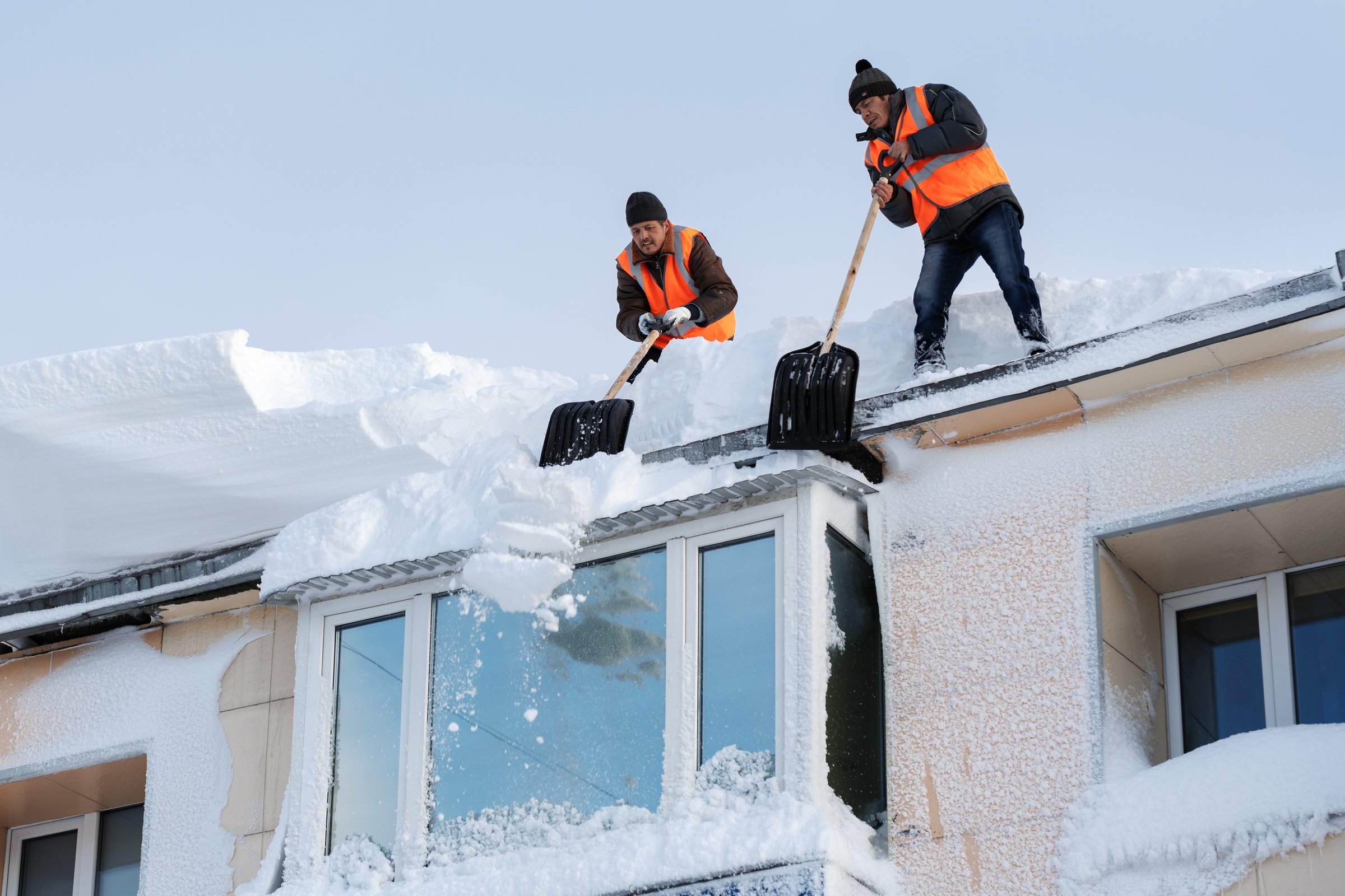 Roof snow removal Anchorage Alaska - two professional workers in safety vests clearing snow from residential roof demonstrating expert snow removal services