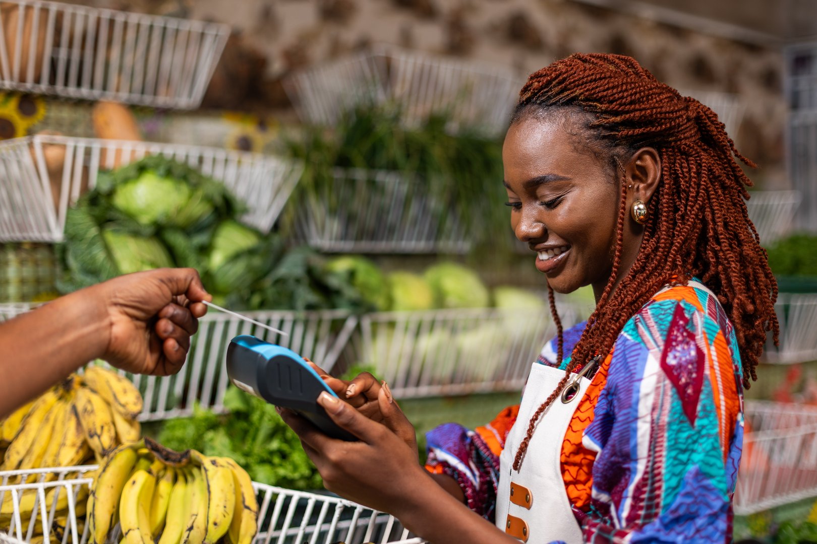 African woman small business owner receives contactless card payment via Point Of sale (POS) terminal, at her fresh produce market where fruits and vegetables are sold.