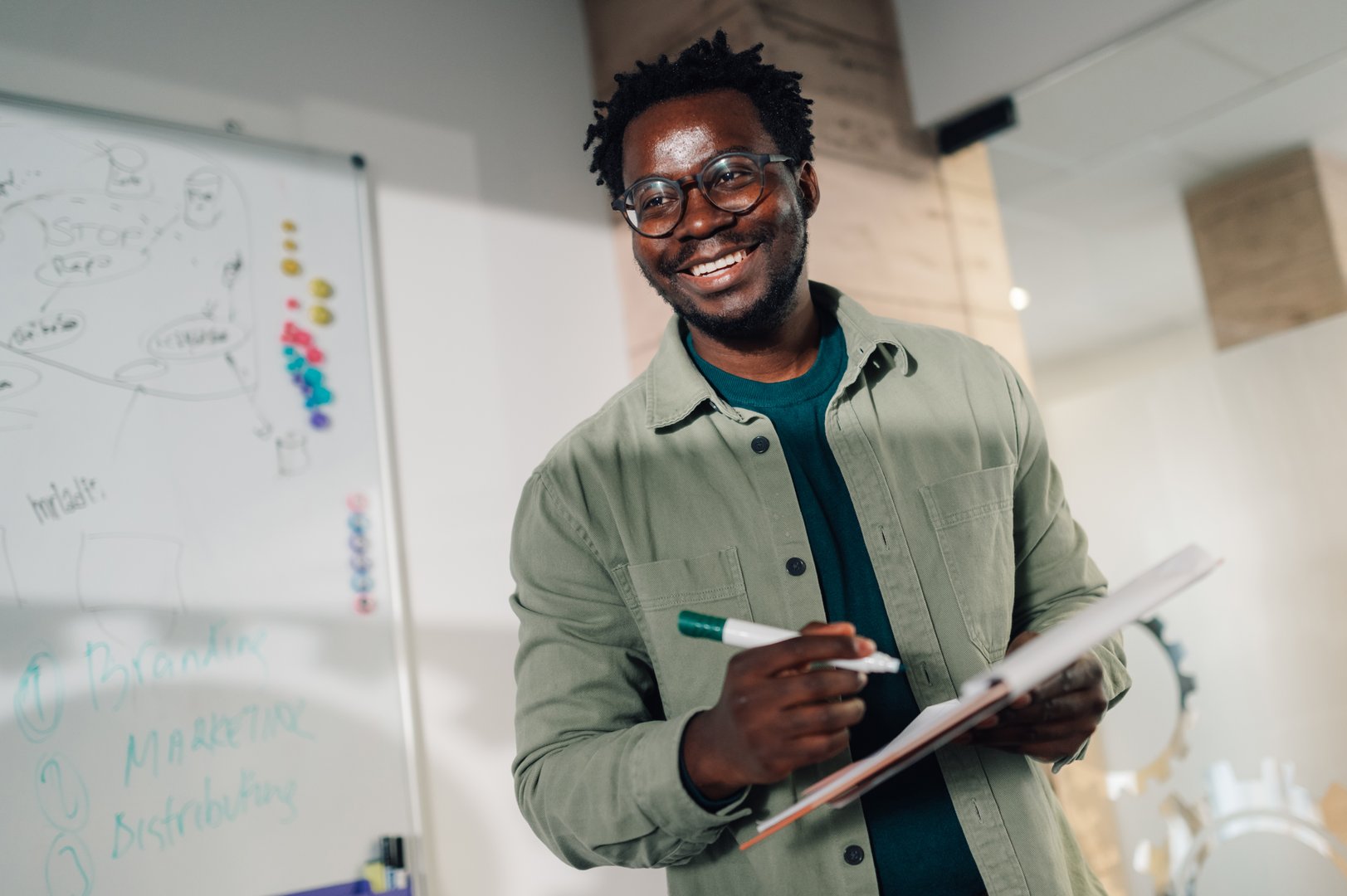 African american businessman happily smiles in a modern meeting room, holding a notebook and marker near a whiteboard with a business plan, exuding confidence and positivity