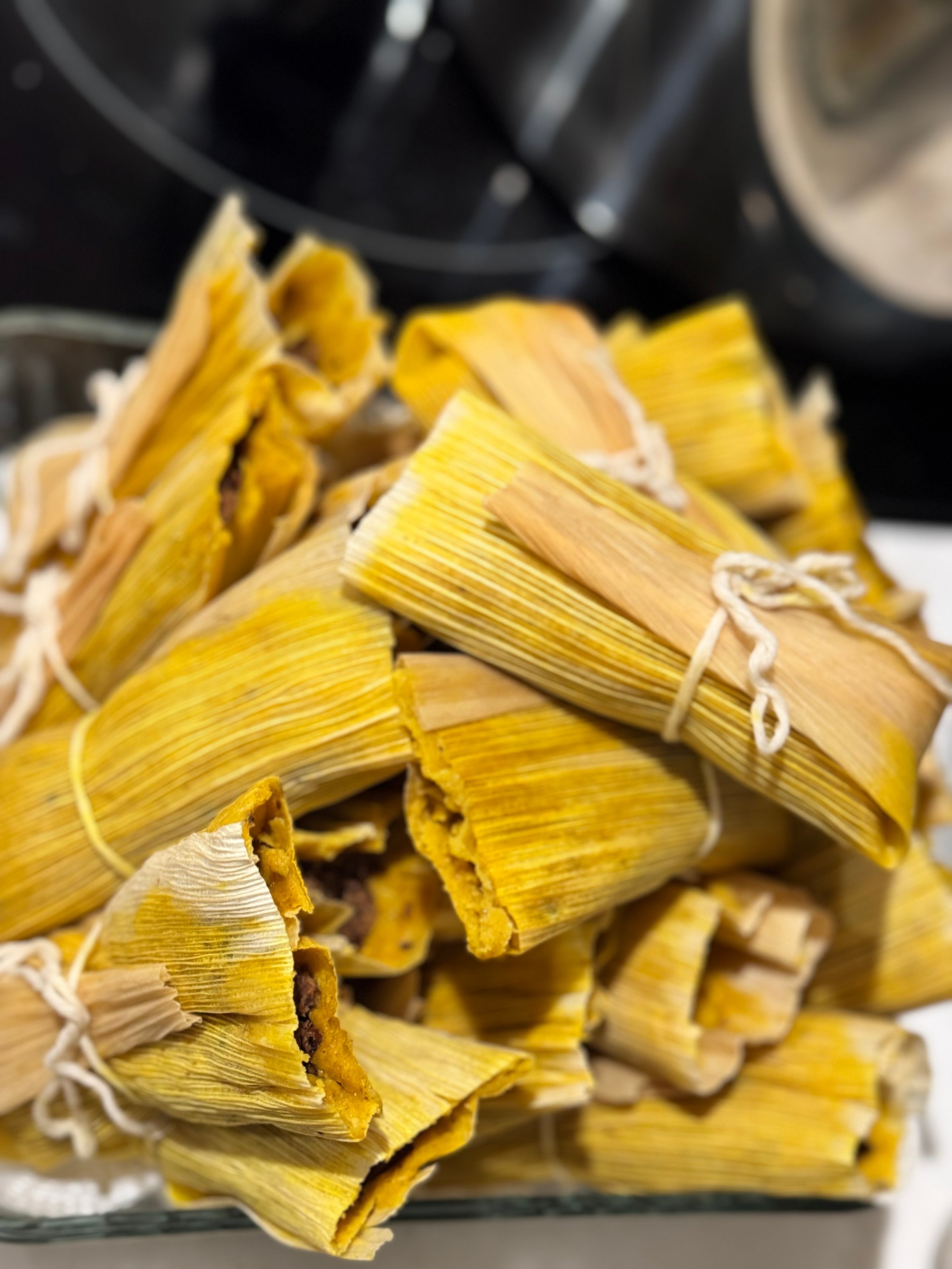 A pile of tamales wrapped in corn husks, some tied with string, displayed on a glass dish.