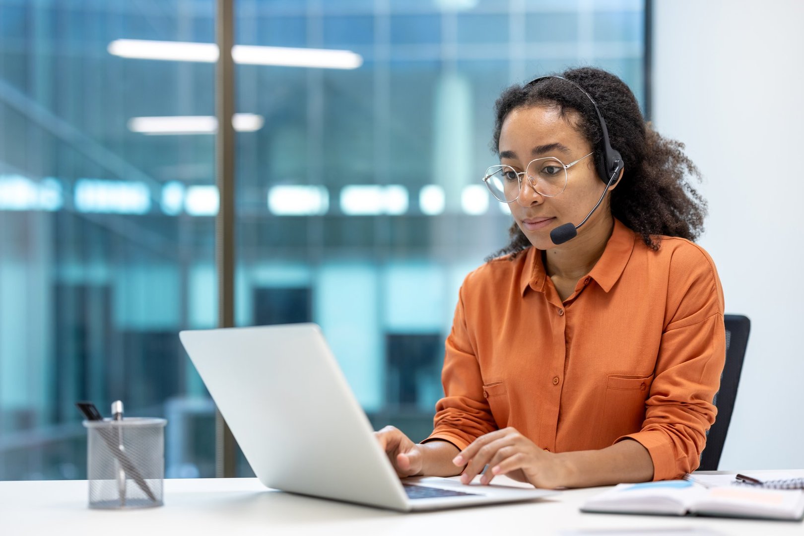 Serious focused woman working with laptop at workplace using headset phone for video call, businesswoman customer service customer support worker inside office.