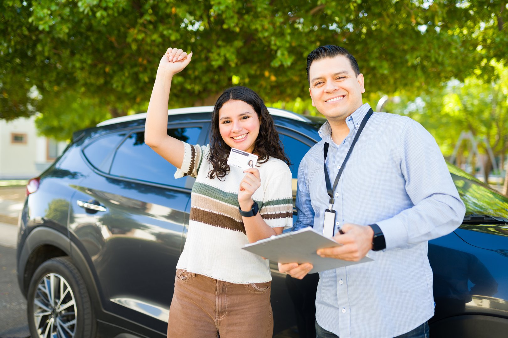 Cheerful young woman celebrates passing her driving test and obtaining her driver's license with her instructor standing next to the car
