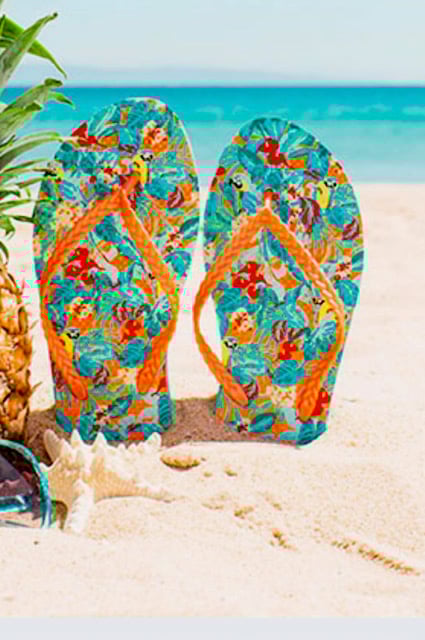 Colorful flip-flops with a tropical print standing upright in the sand on a beach, near sunglasses and a starfish.