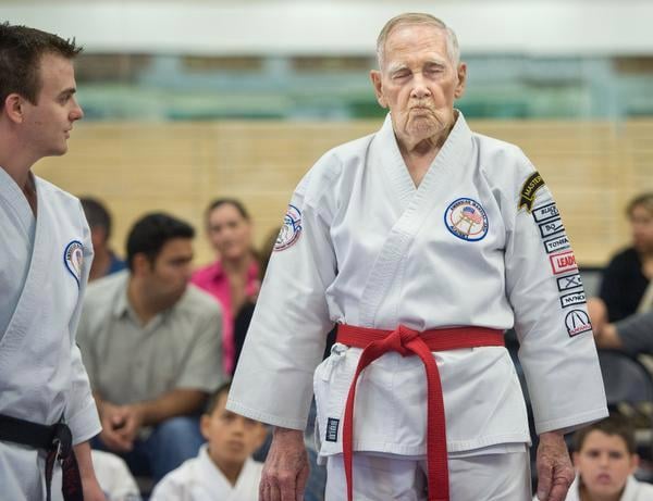 An elderly martial artist in a white gi with a red belt stands in a dojo, surrounded by other participants and spectators.