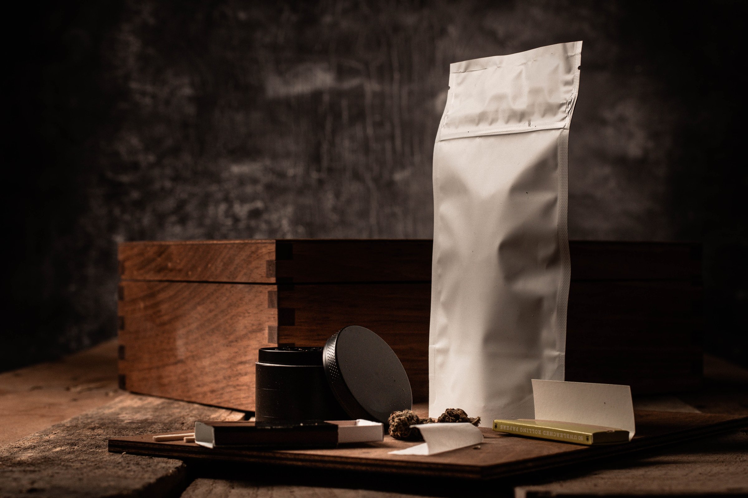 A generic package of cannabis with a grinder, rolling papers, matches, and loose cannabis on a wooden board.