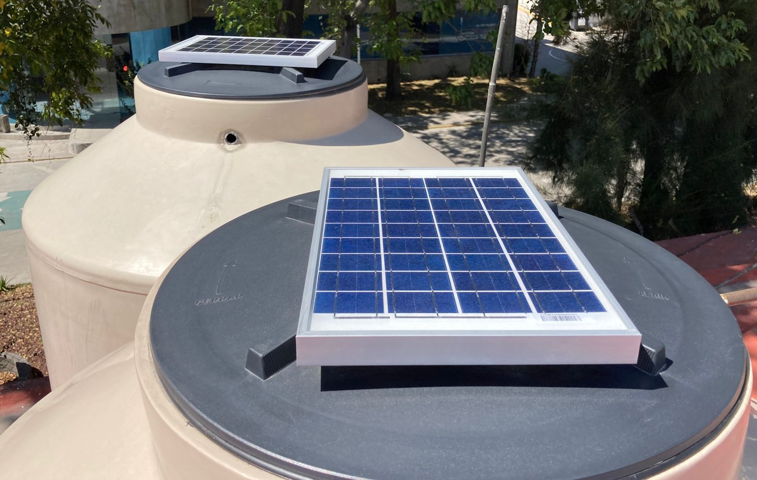 Solar panels installed on top of two large water tanks outdoors, surrounded by trees and a building in the background.