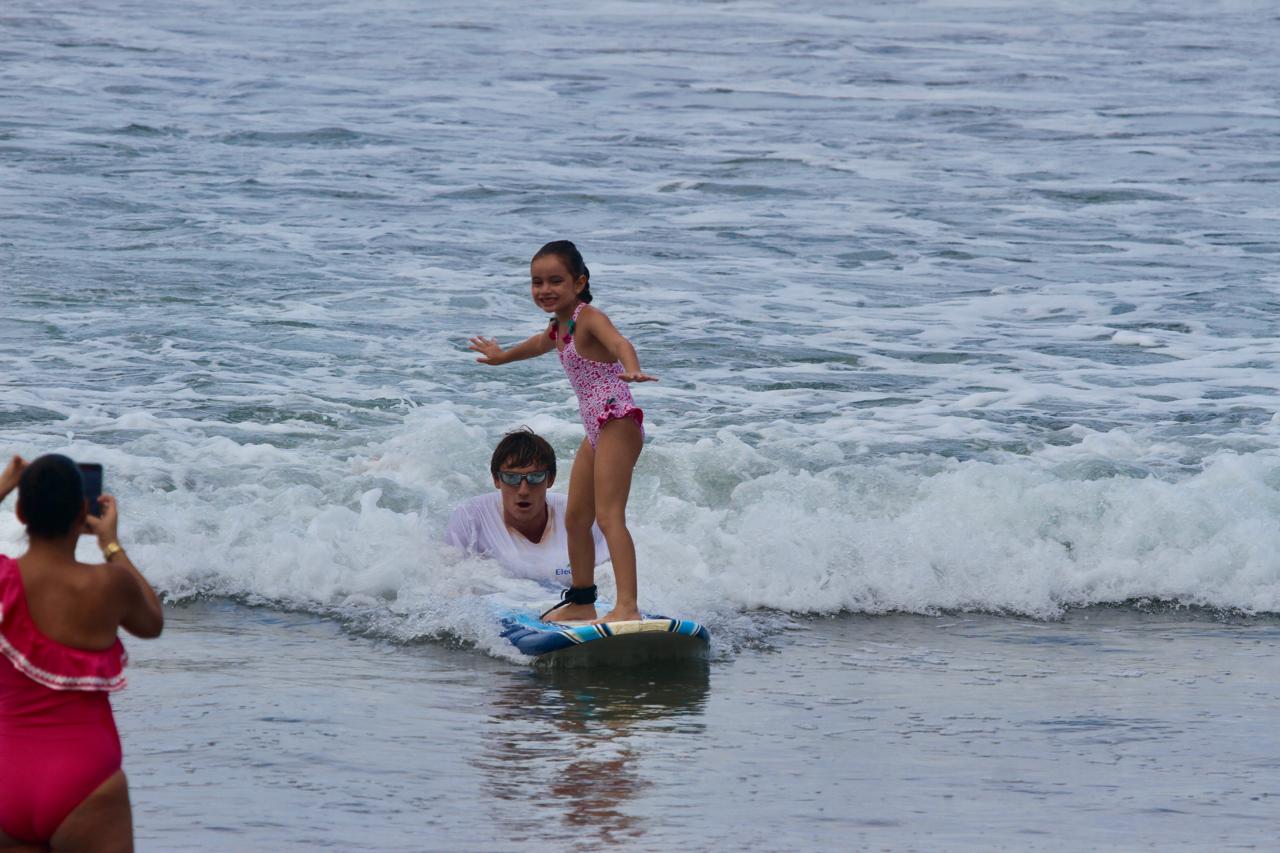 A young girl in a pink swimsuit stands on a surfboard riding a wave, while a person in a red swimsuit takes her photo.