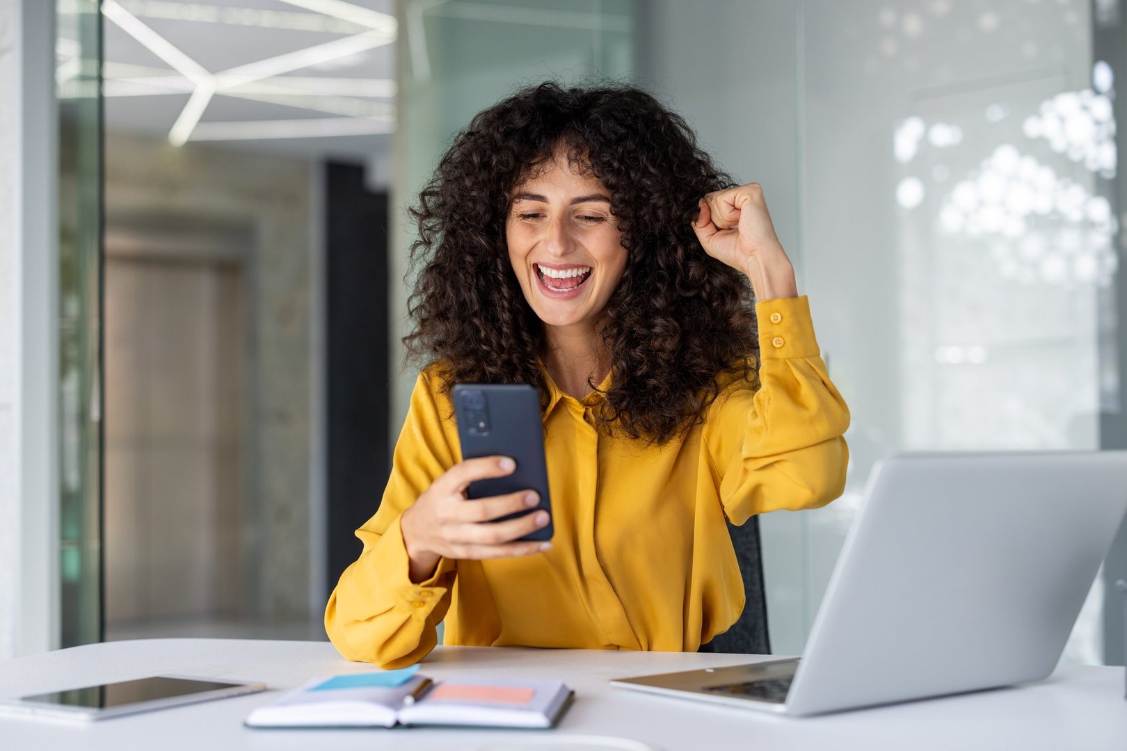 Latin American businesswoman celebrates success in office holding smartphone, displaying excitement. Laptop on desk, showing workplace environment. Emotion of achievement, using technology for communication.