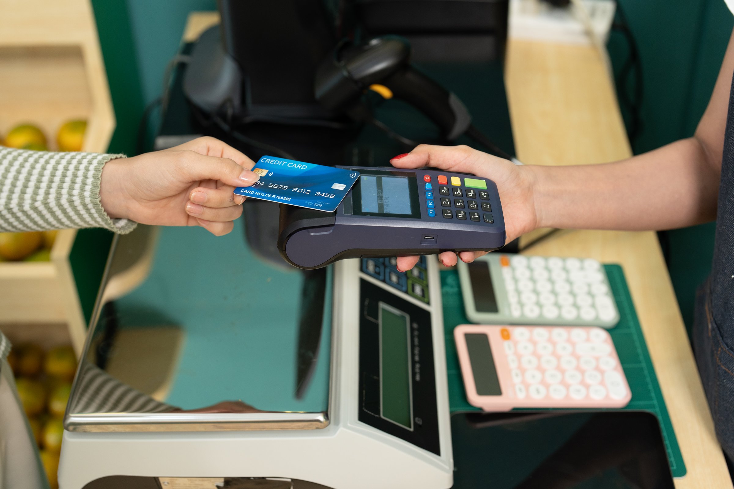 Caucasian adult female paying for fruit at small business fruit shop using contactless credit card payment method by tapping card on terminal held by female Asian fruit shop cashier in apron.