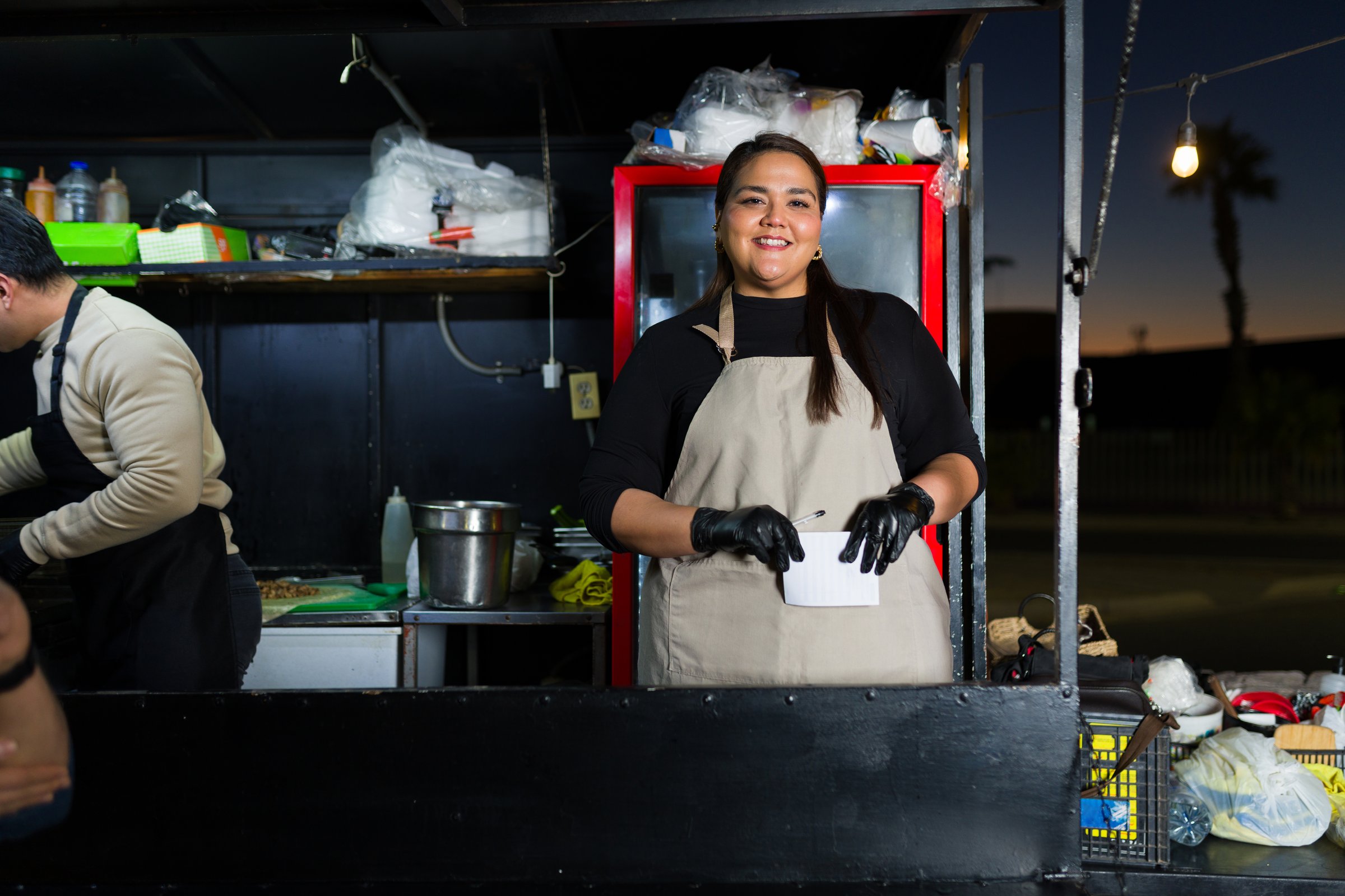 Plus size latin american woman wearing gloves and apron working in a food truck at night, taking orders and smiling
