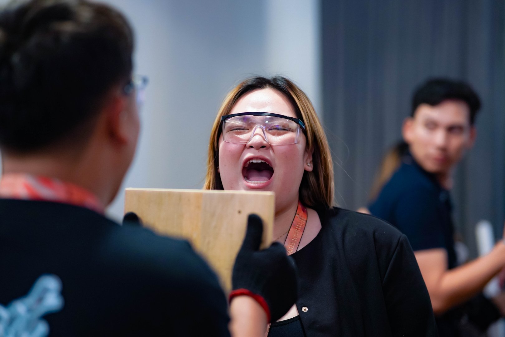 Woman wearing safety goggles shouting at a wooden board held by another person, with a third person blurred in the background.