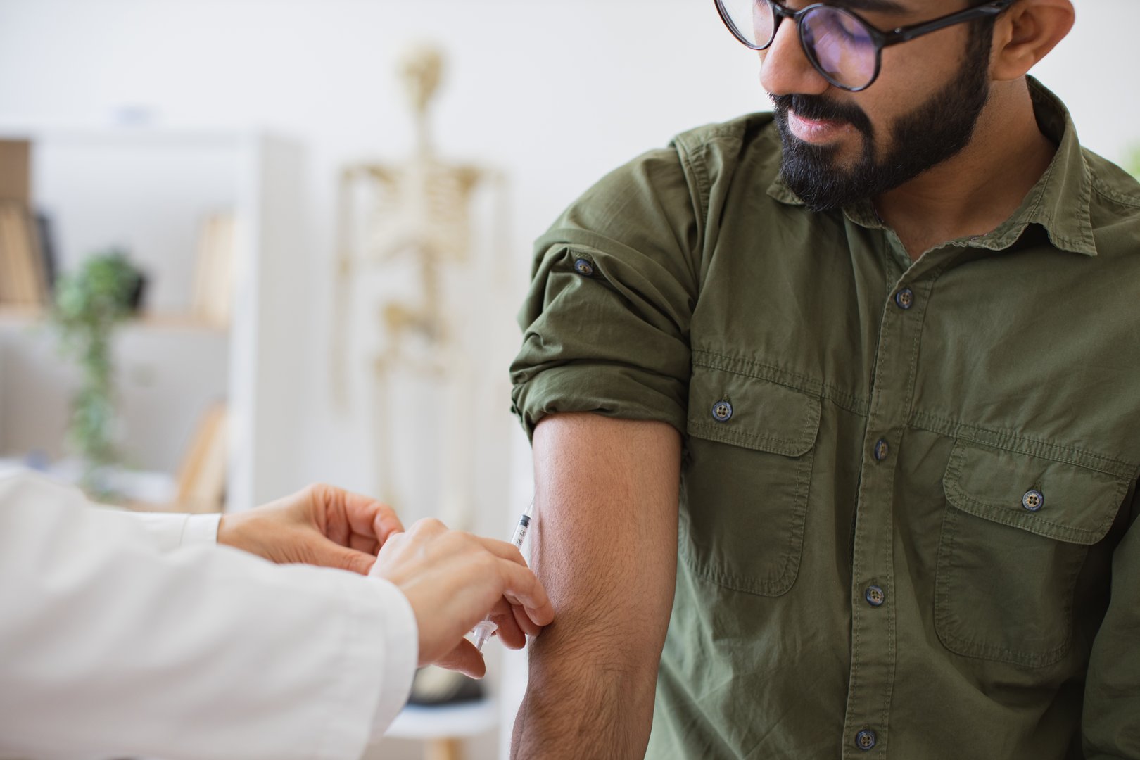 Cropped view of hand of general practitioner giving injection to boost immunity for bearded patient. Doctor in lab coat holding syringe with seasonal flu vaccine while young man sitting in exam room.