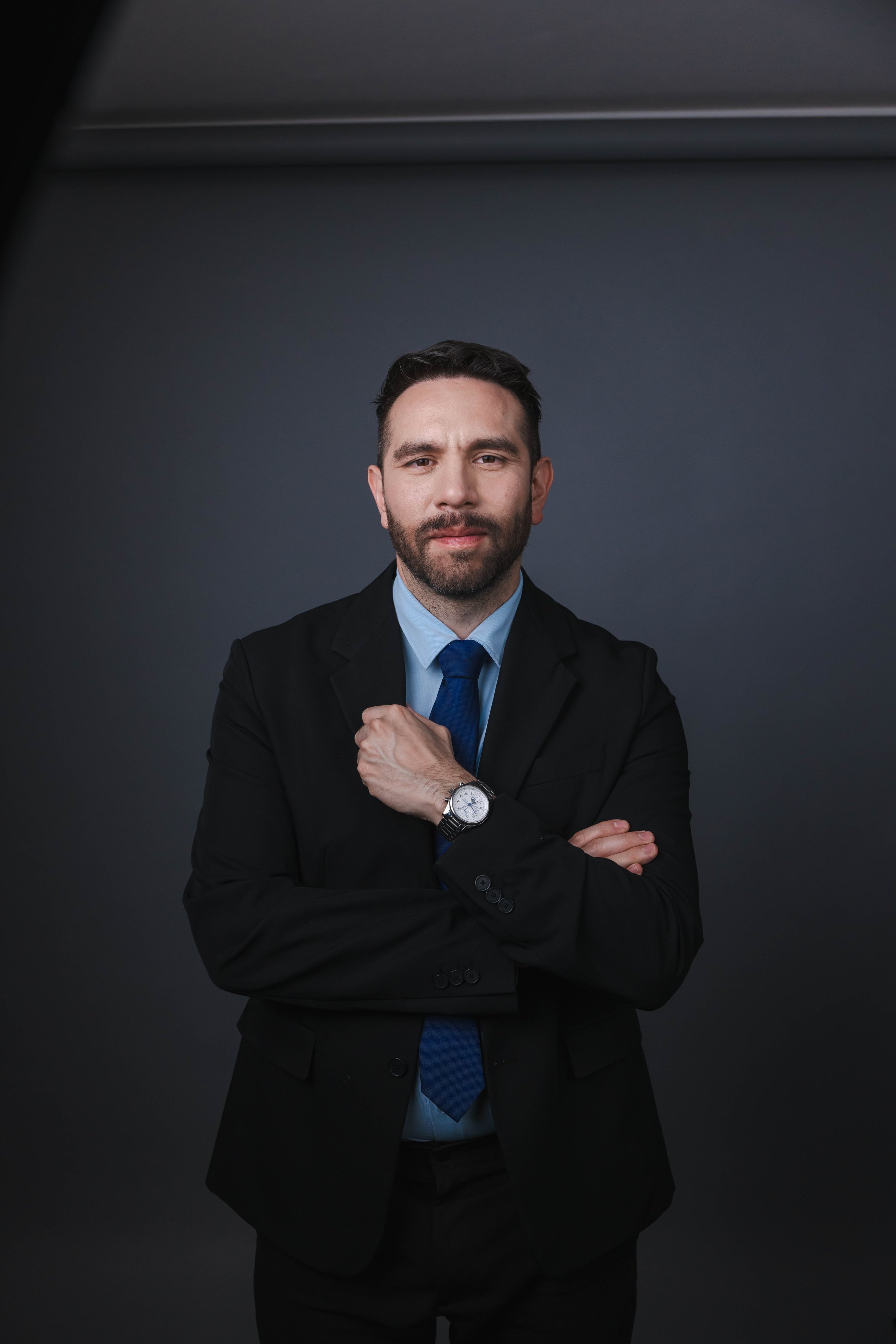 Man in a suit and tie stands confidently with arms crossed against a dark background.