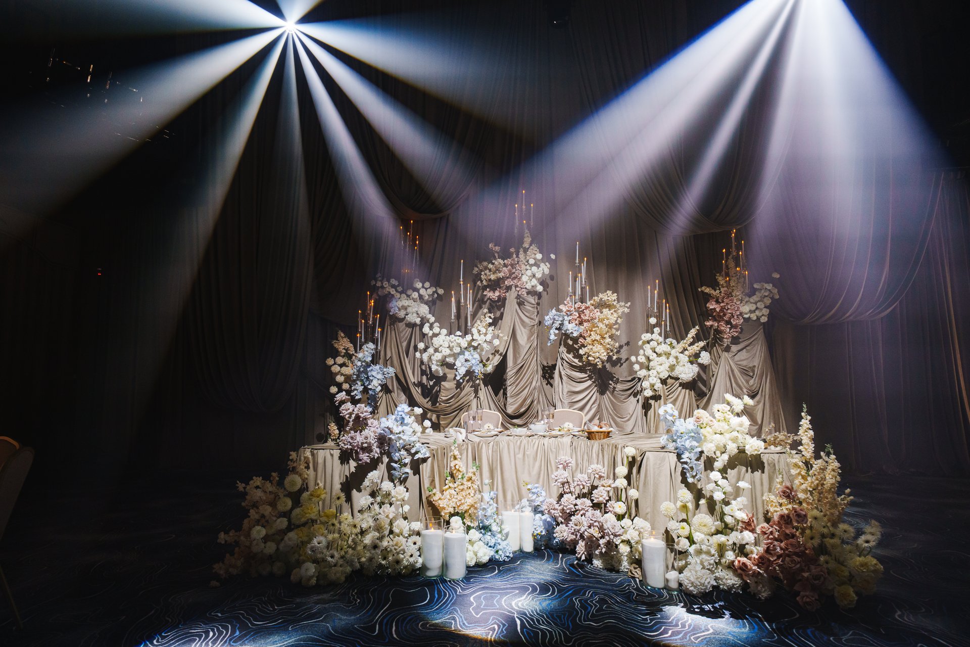 A lavish arrangement of flowers and candles fills the venue, illuminated by soft beams of light. This stunning setup creates a romantic atmosphere for an unforgettable gathering.