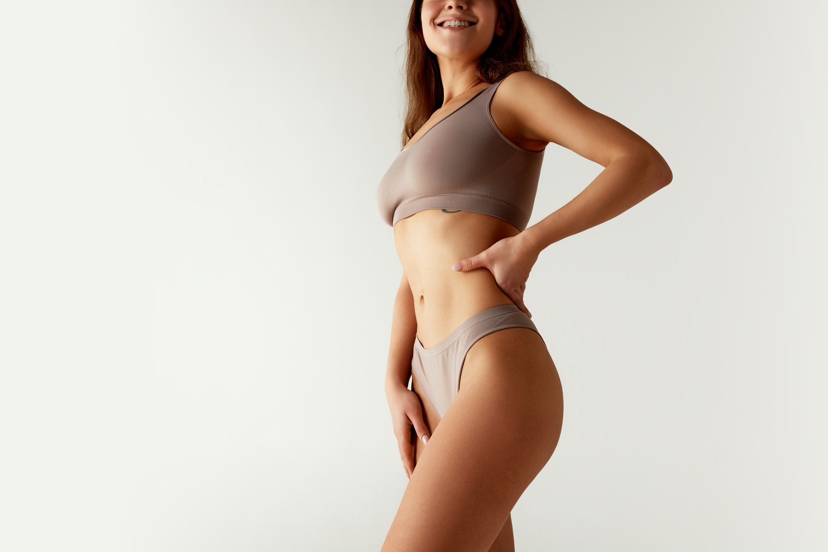 Cropped portrait of woman in sports bra and underwear set, confidently placing hands on her hips against grey studio background. Concept of beauty treatments, dieting, female health, spa procedures.