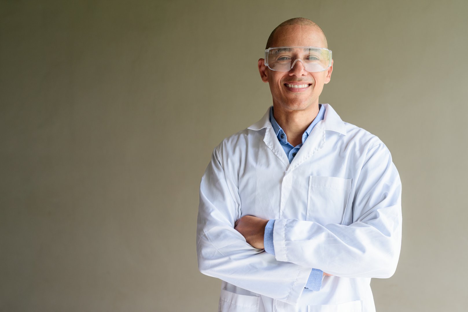 Confident male scientist in white lab coat and protective glasses posing against plain wall. Studio science concept.