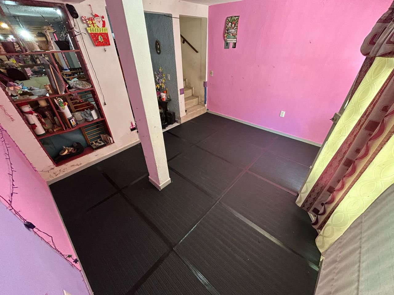 Room with black foam flooring, pink walls, and a shelf with items. Curtains and a staircase are visible in the background.