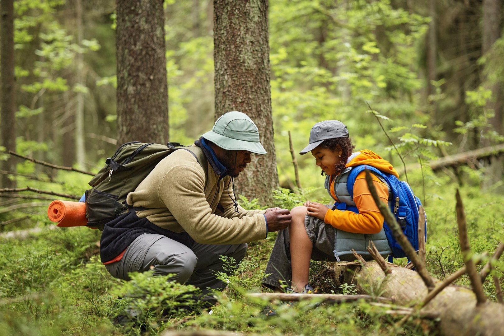 Black middle aged man kneeling in forest helping Black child with injured knee during hiking trip, both wearing backpacks and hats, surrounded by green trees and foliage
