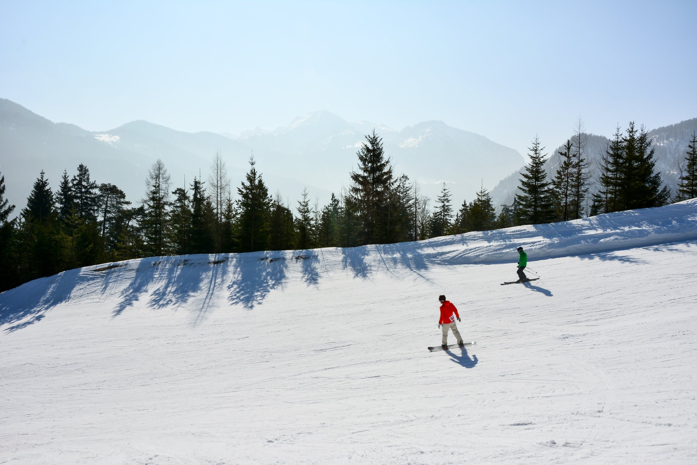 A skateboarder and a skier of an unrecognizable appearance are going down the ski track. Active winter recreation at a ski resort against the background of mountains and the sky