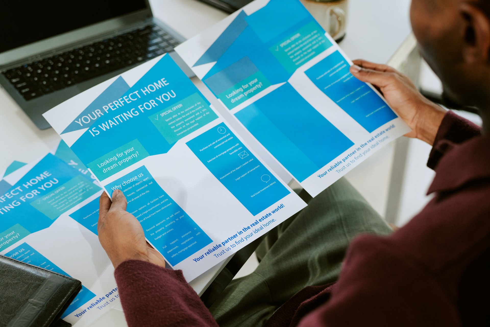 Black man sitting at desk holding real estate brochure with blue graphics, reading information about home buying options, partial view of hands and arms visible, office setting