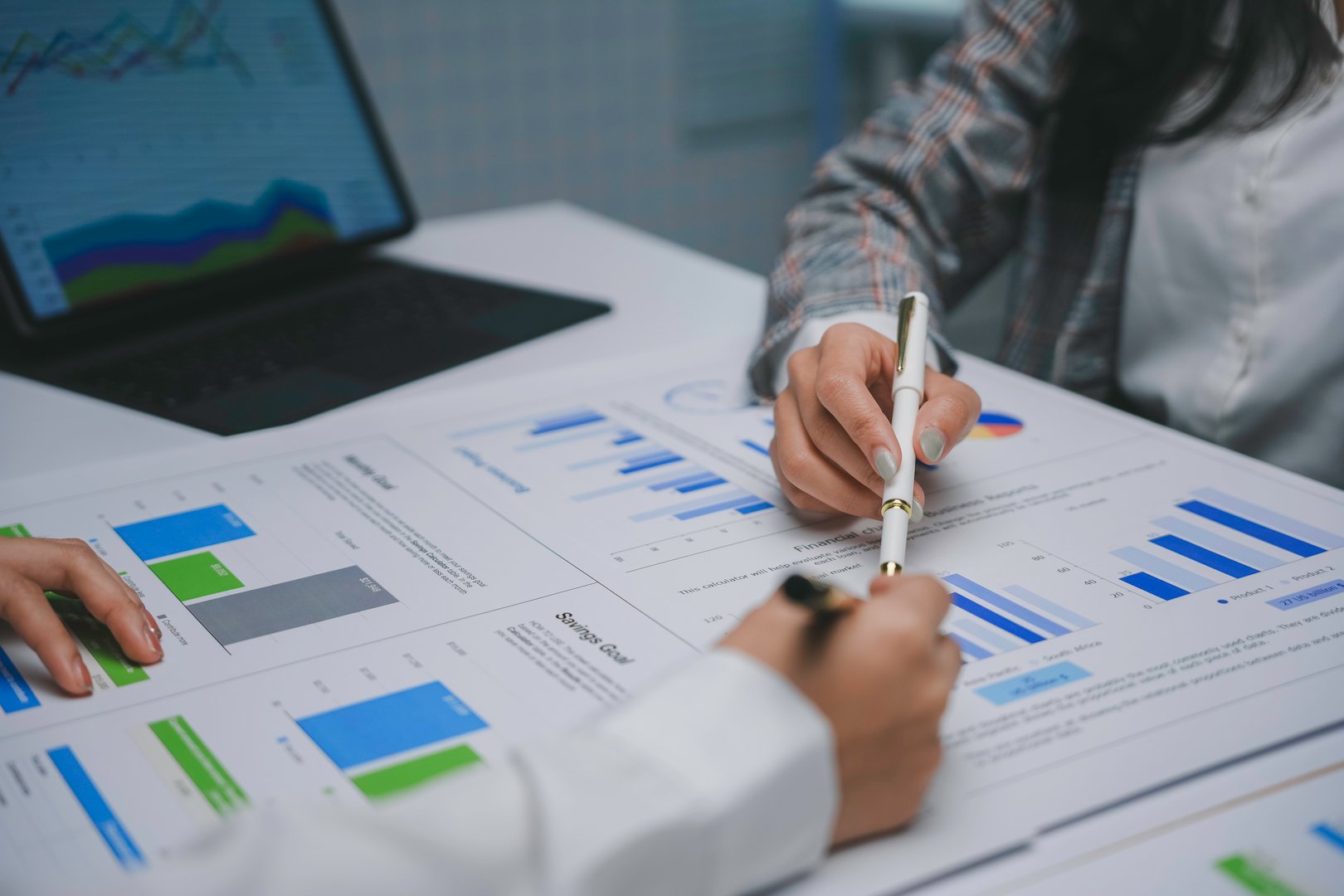 Two businesswomen are pointing at financial charts and graphs printed on paper, discussing company strategy using a laptop and tablet, analyzing business growth and planning investments