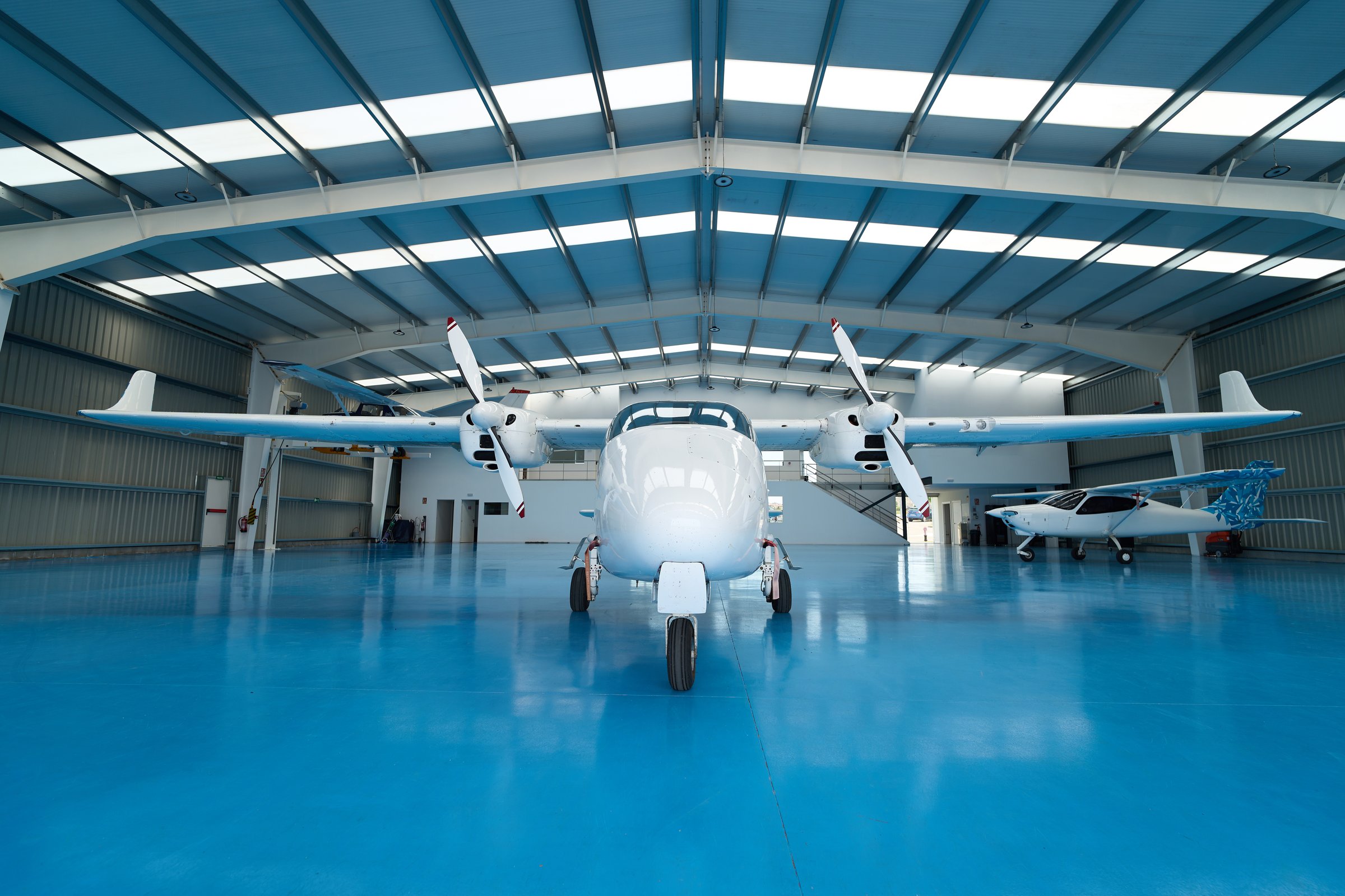 White twin-engine aircraft parked prominently in a hangar with a blue floor, possibly at an aviation school