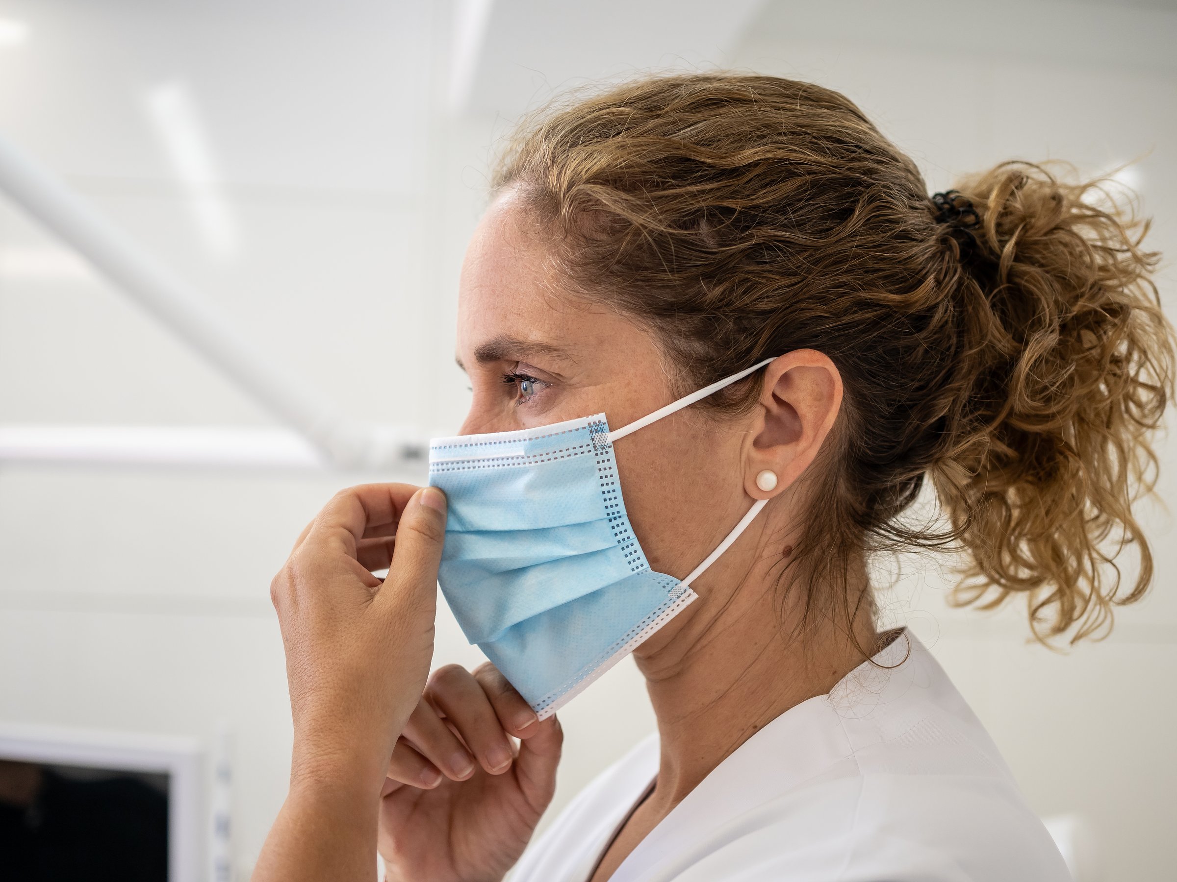 A healthcare worker adjusts a light blue mask while in a clinical environment, demonstrating proper face covering practices for safety and health.