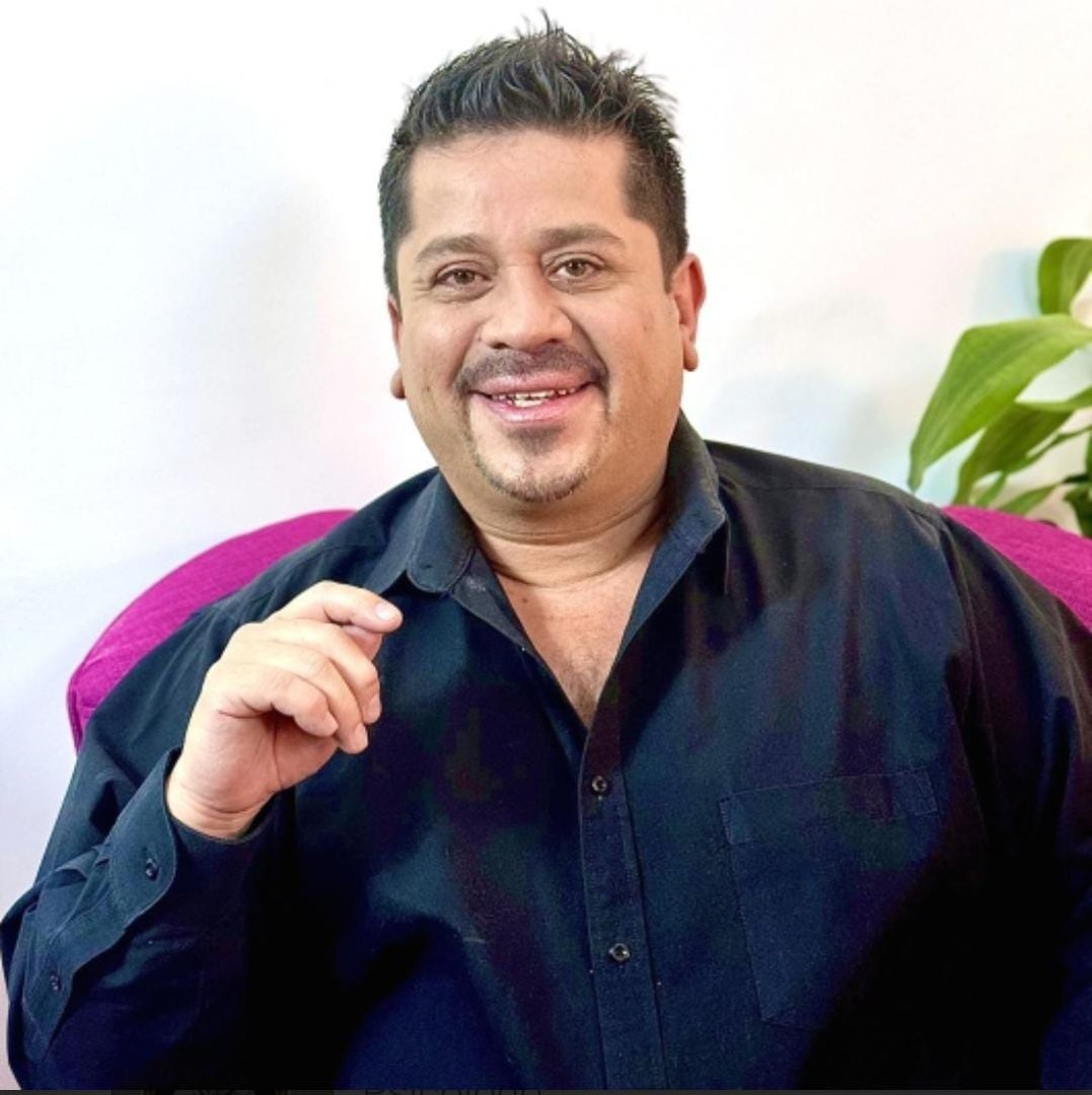 Man in a black shirt smiling, sitting indoors, with a plant in the background.