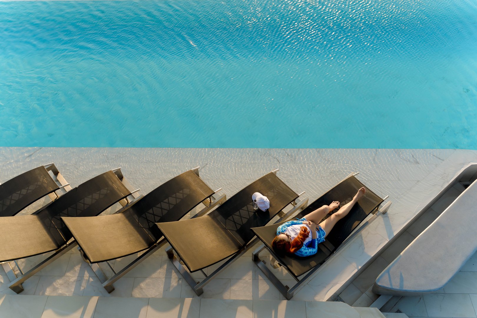 Top view of young woman relaxing alone on a lounge chair by rooftop infinity pool in bright sunlight, enjoying peaceful summer moment and clear blue water