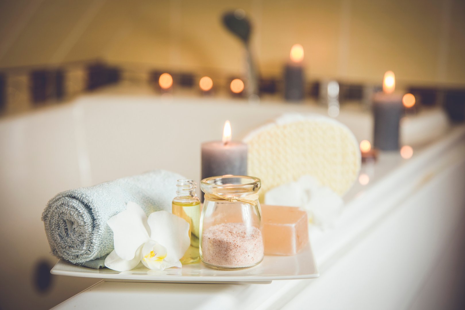 White ceramic tray with home spa supplies in home bathroom for relaxing rituals. Candlelight, salt soap bar, bath salt in jar, massage, bath oil in bottle, blue rolled towel, natural sponge.