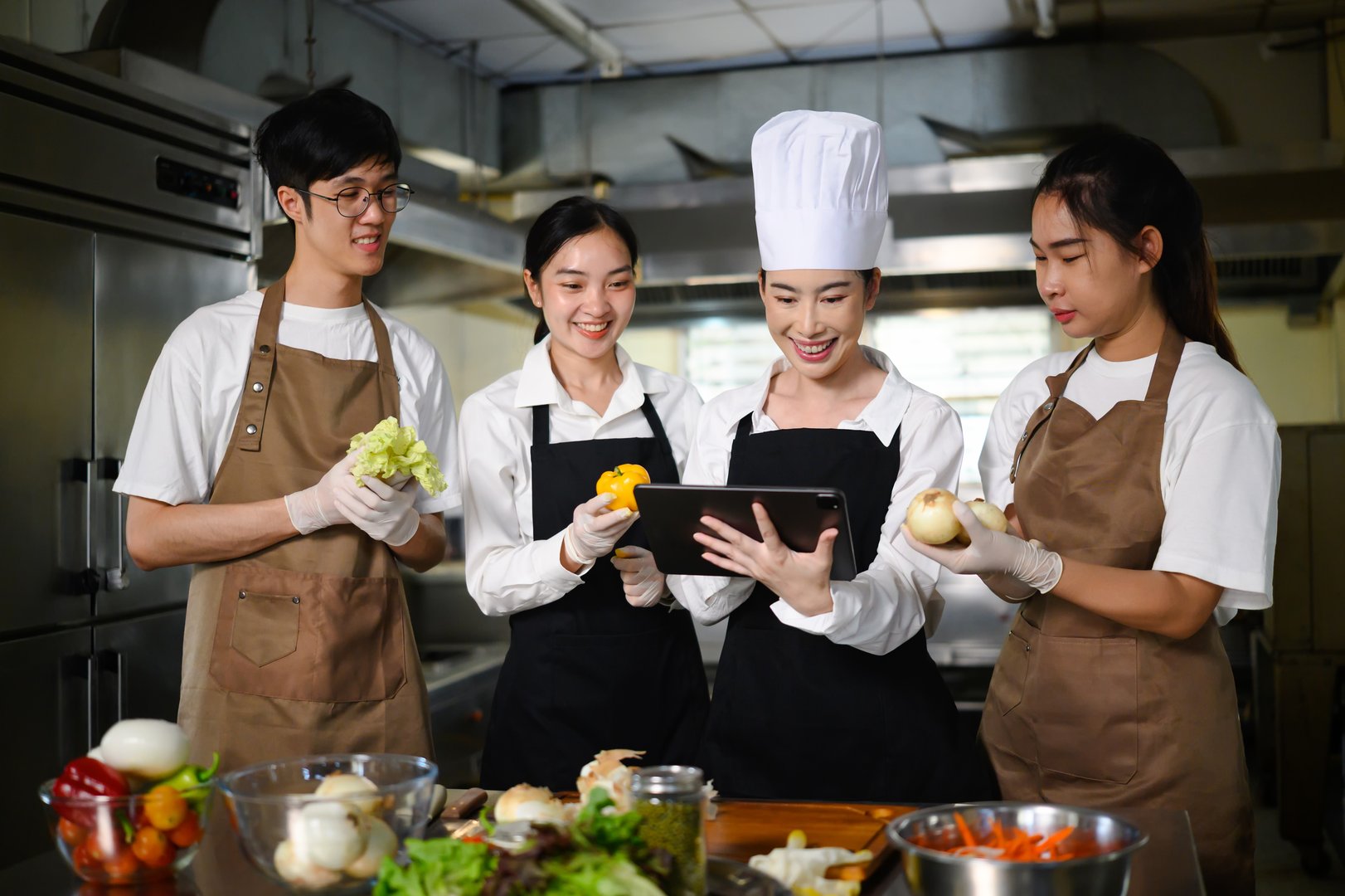 Smiling chef teacher using a tablet to explain a recipe to students in a professional kitchen.